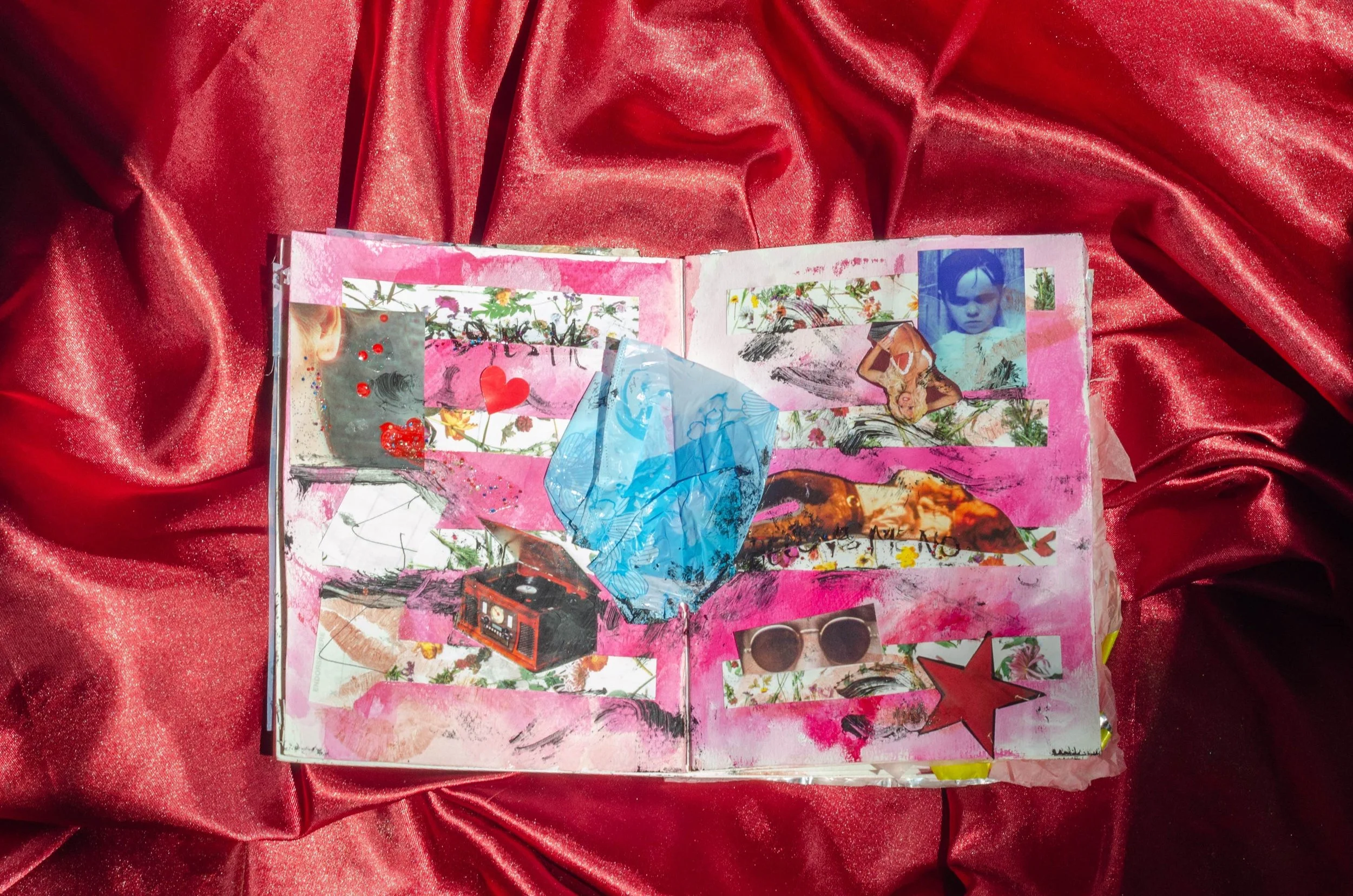 An art journal with mixed media collage elements on a pink background, featuring photos, drawings, and embellishments, placed on a red satin fabric.