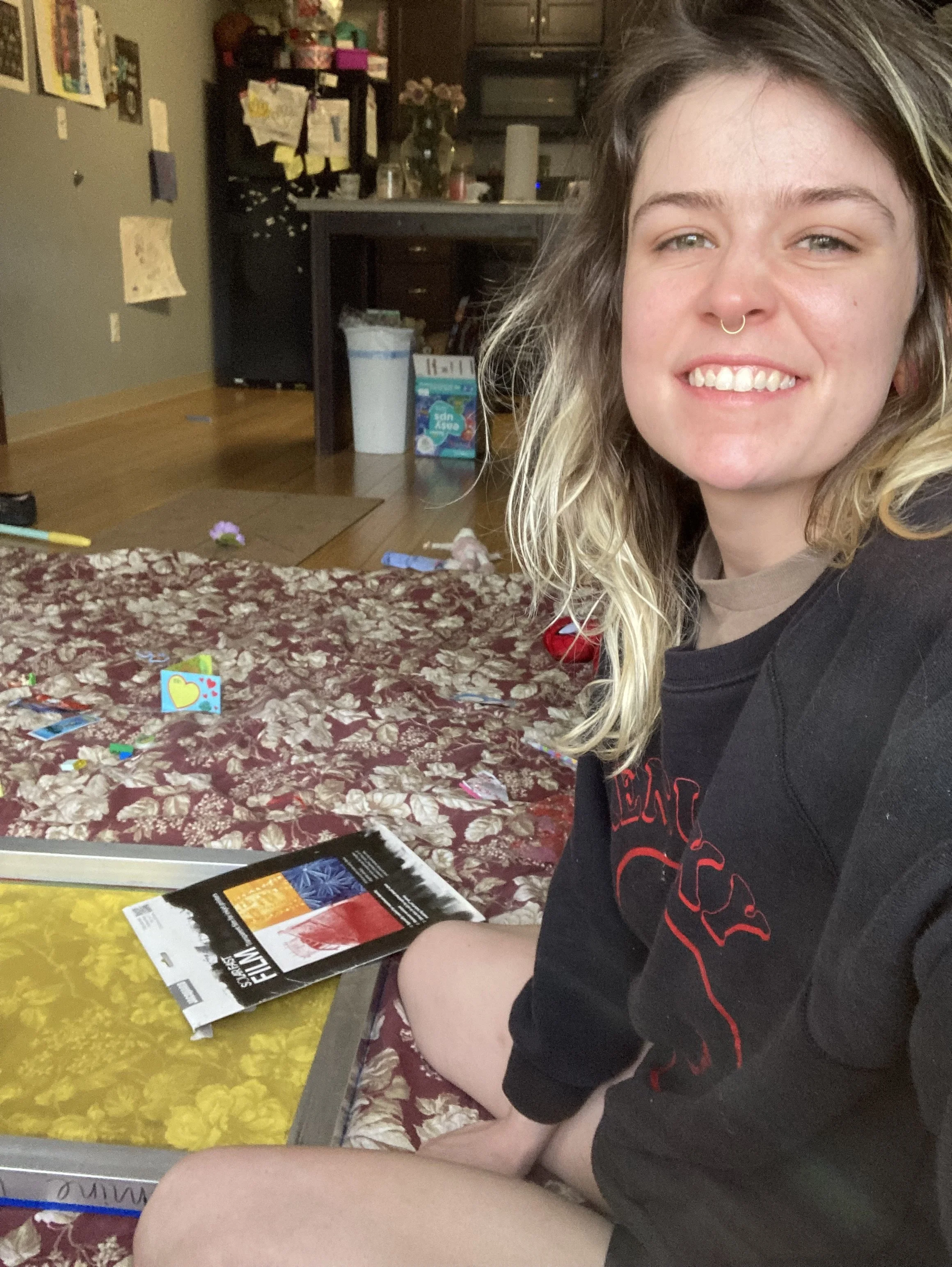 A woman sitting on a bed with a patterned blanket, smiling at the camera, with a magazine in front of her. The background shows a cluttered kitchen and living area.