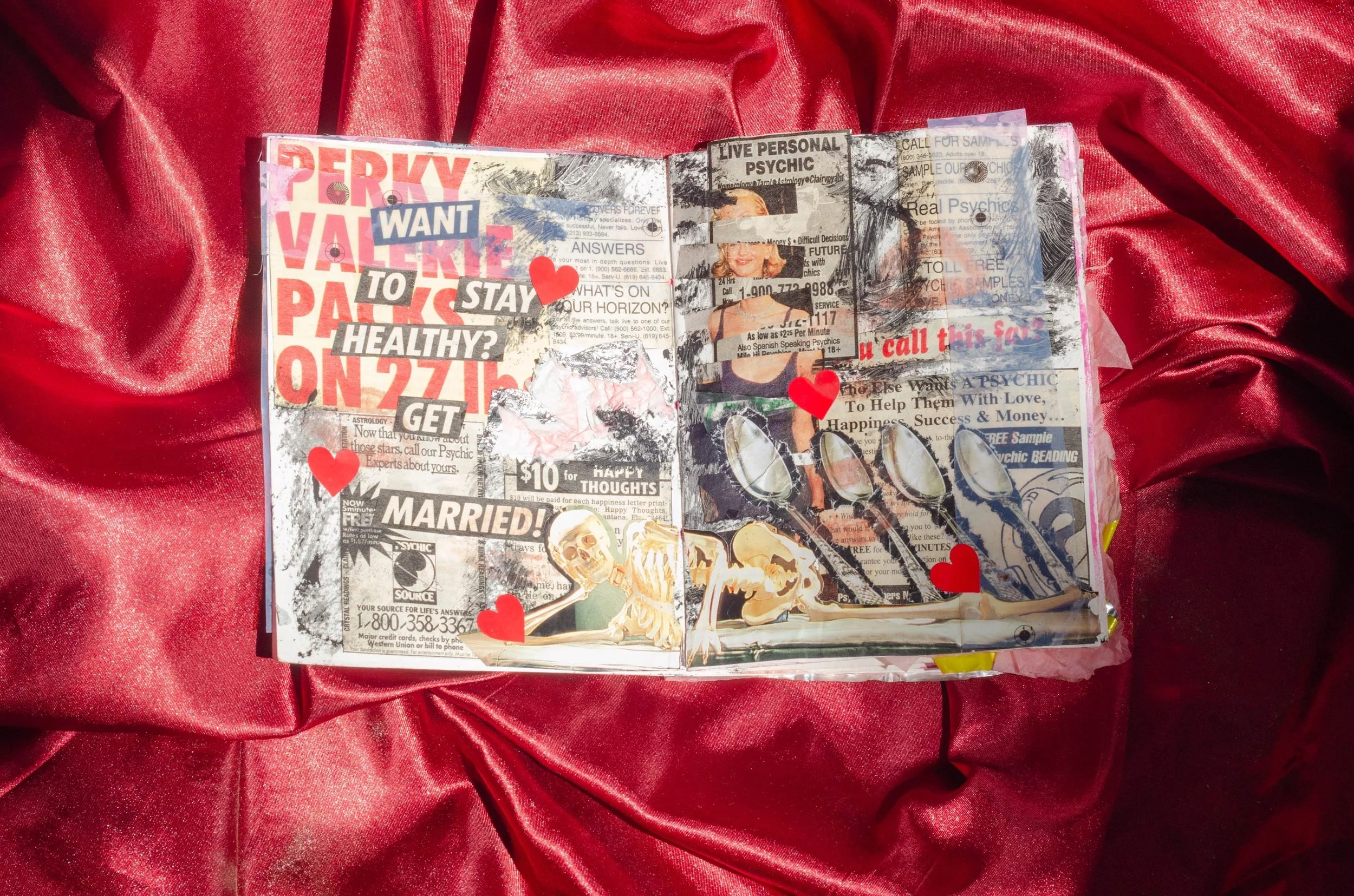 An open mixed-media collage art journal on red satin fabric, featuring black-and-white newspaper clippings, colorful stickers, and cutouts with religious and romantic themes, including skeleton illustrations and red heart shapes.