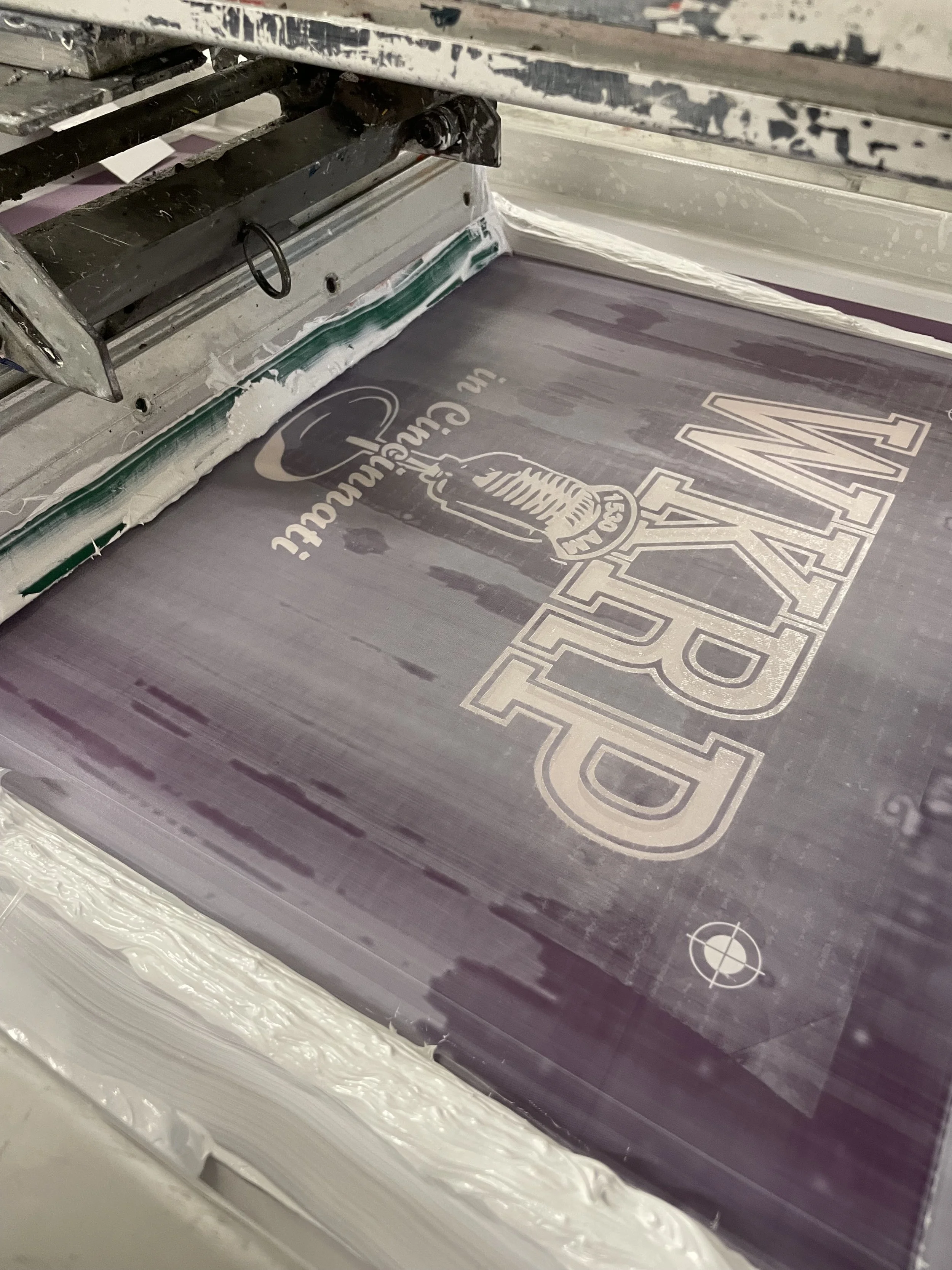 Silkscreen printing process creating a logo on a sheet of material. A partially printed design with text and graphics is being transferred through a screen.
