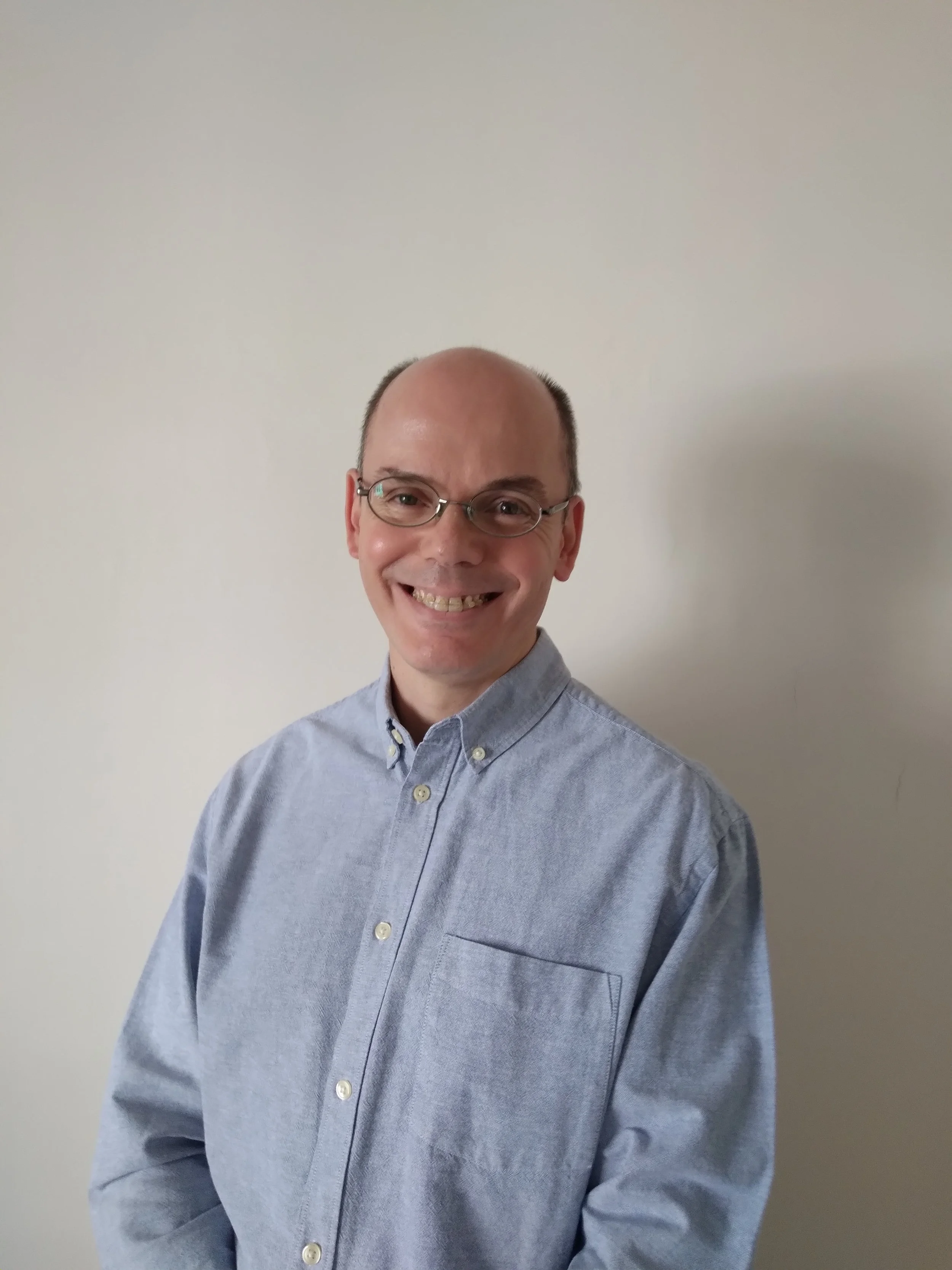 Hombre de camisa azul y gafas sonriendo frente a pared blanca.