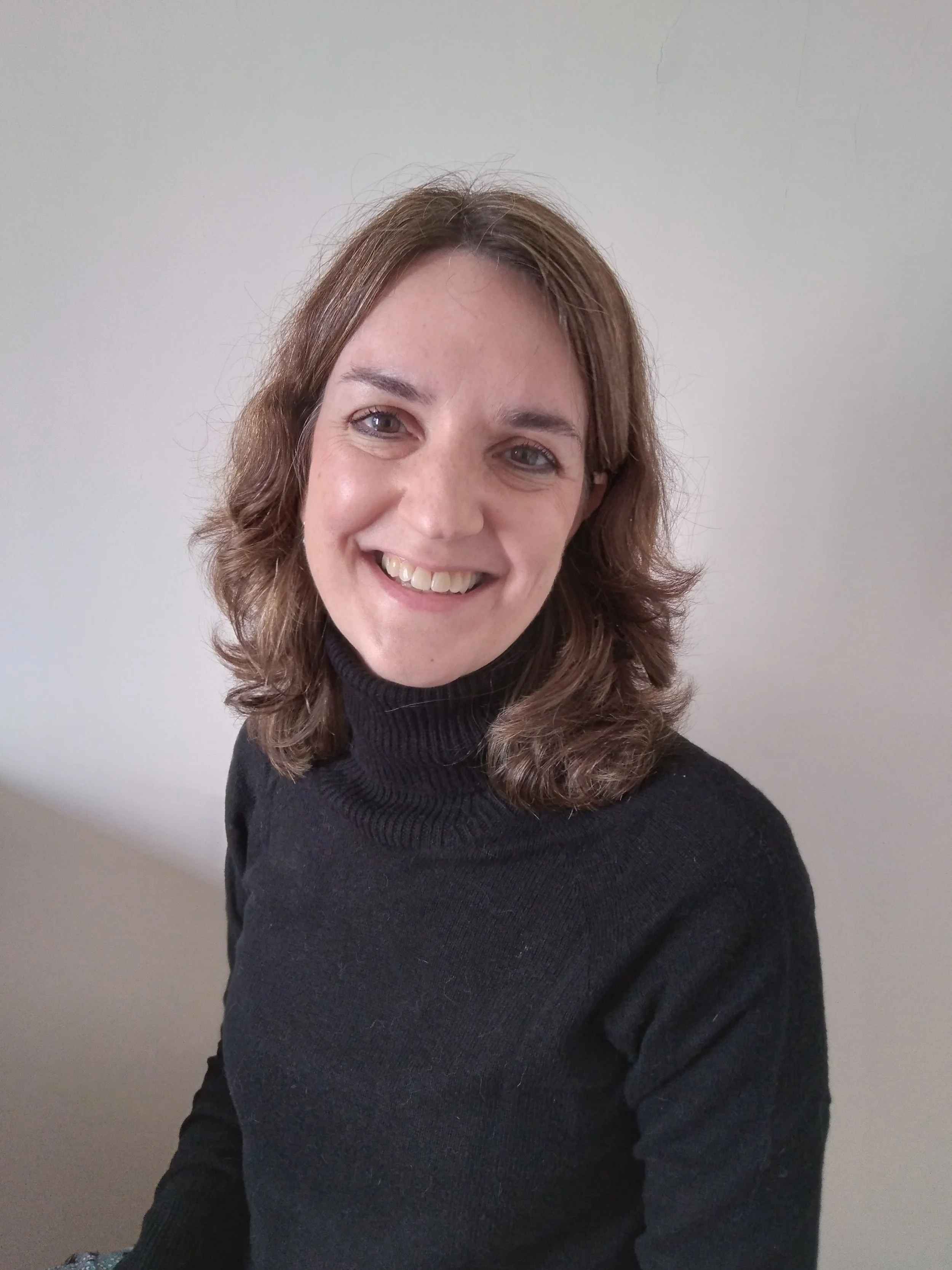 Retrato de una mujer sonriendo con cabello castaño y rizado, lleva un suéter negro de cuello alto, posa frente a una pared clara.