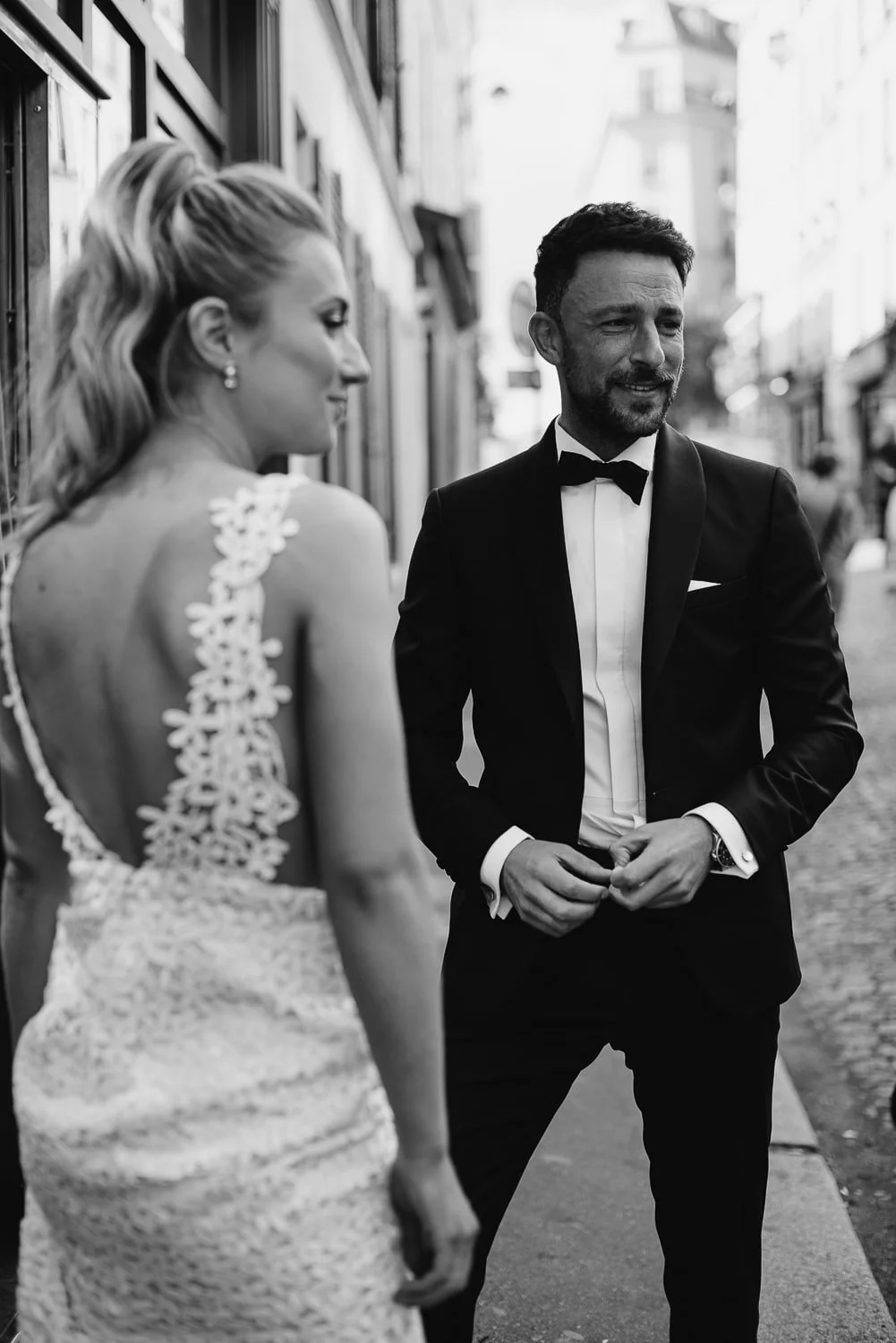 Black and white photo of a bride in a lace dress and a groom in a tuxedo standing on a city street.