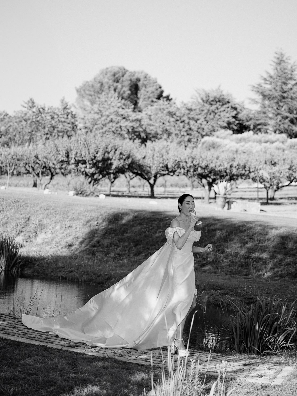 Bride in a flowing gown walking near a small pond, outdoor setting with trees in the background.