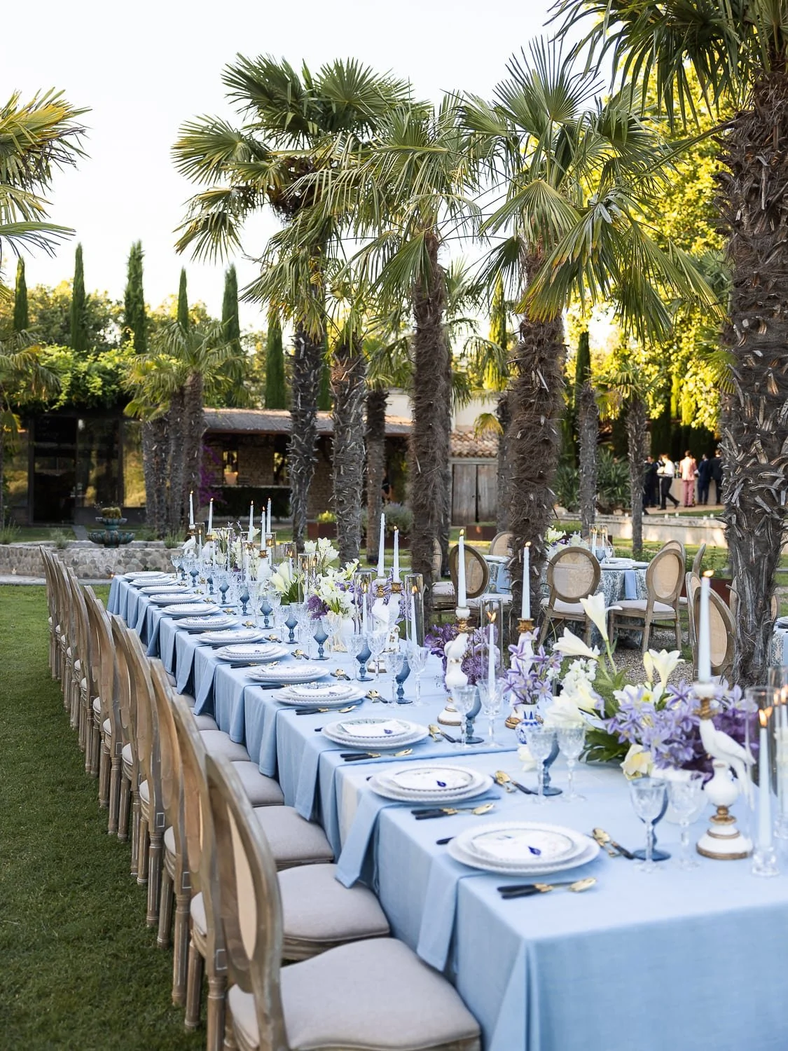 Outdoor dining setup with long tables covered in light blue tablecloths, surrounded by chairs, and decorated with white and purple floral arrangements and candles. Palm trees and greenery are in the background.