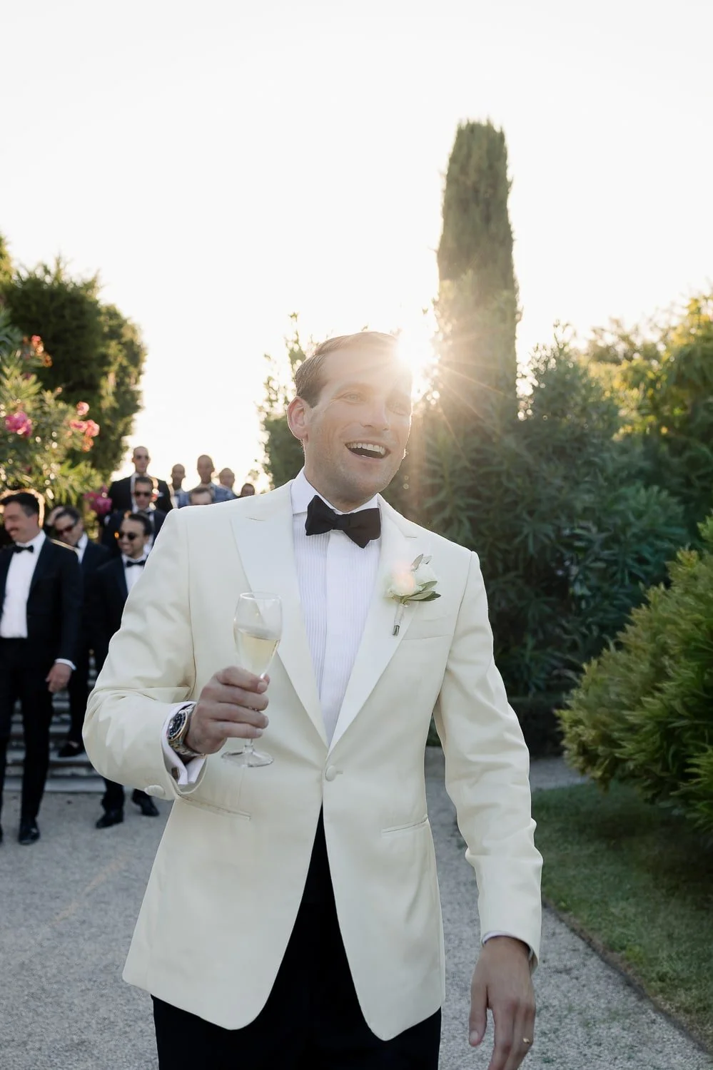 A man in a white tuxedo with a black bow tie, holding a glass of champagne, smiling at a wedding or formal event outdoors in the late afternoon or early evening sunlight.