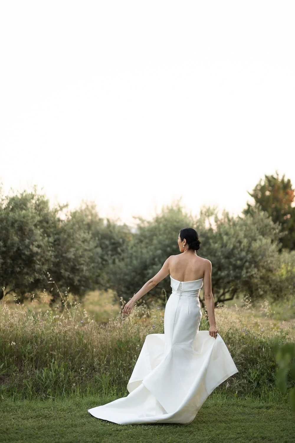 A woman in a white strapless wedding dress walking through a grassy field with trees in the background during sunset.