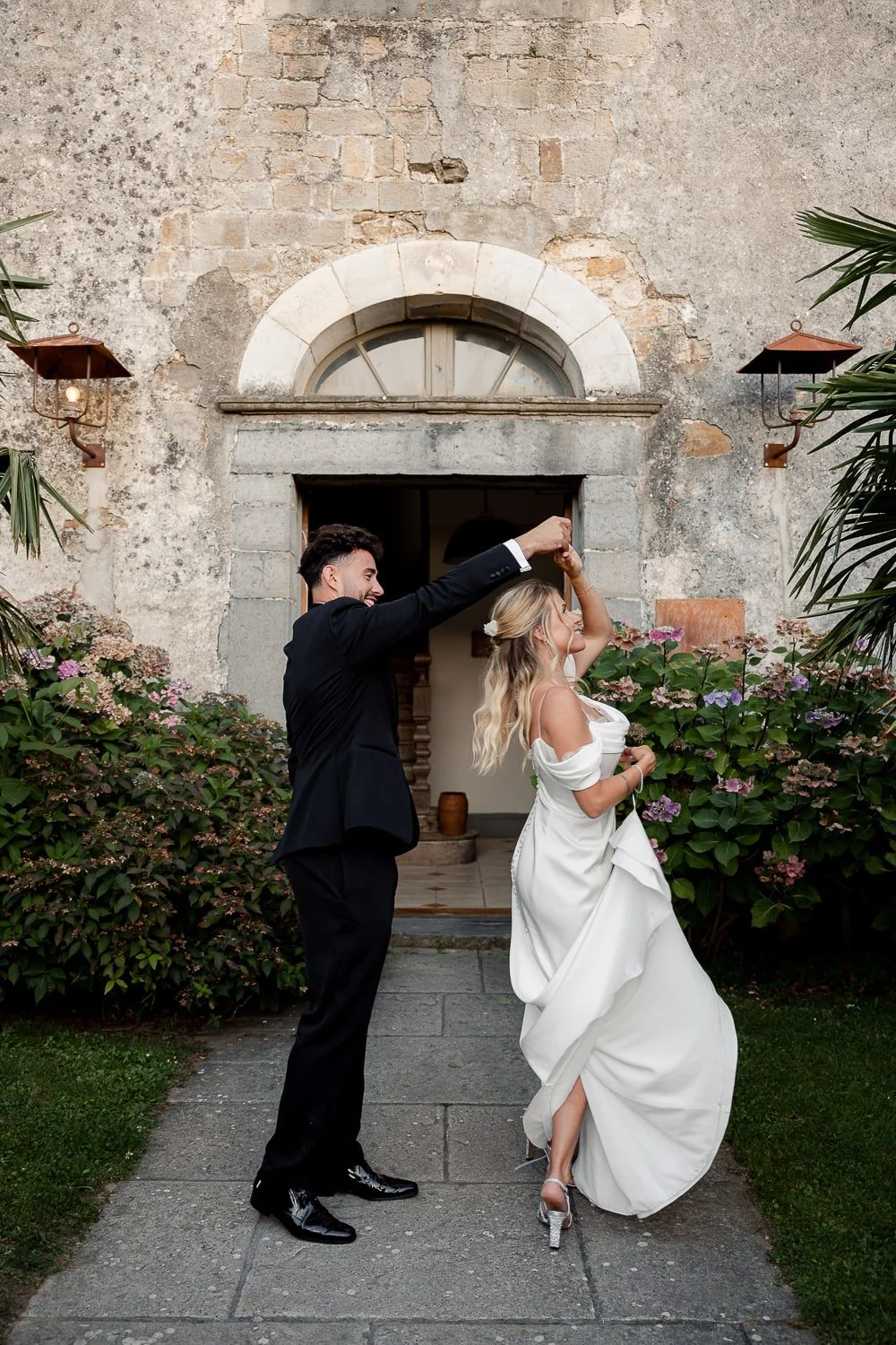 Bride and groom dancing outside rustic stone building with hydrangeas.