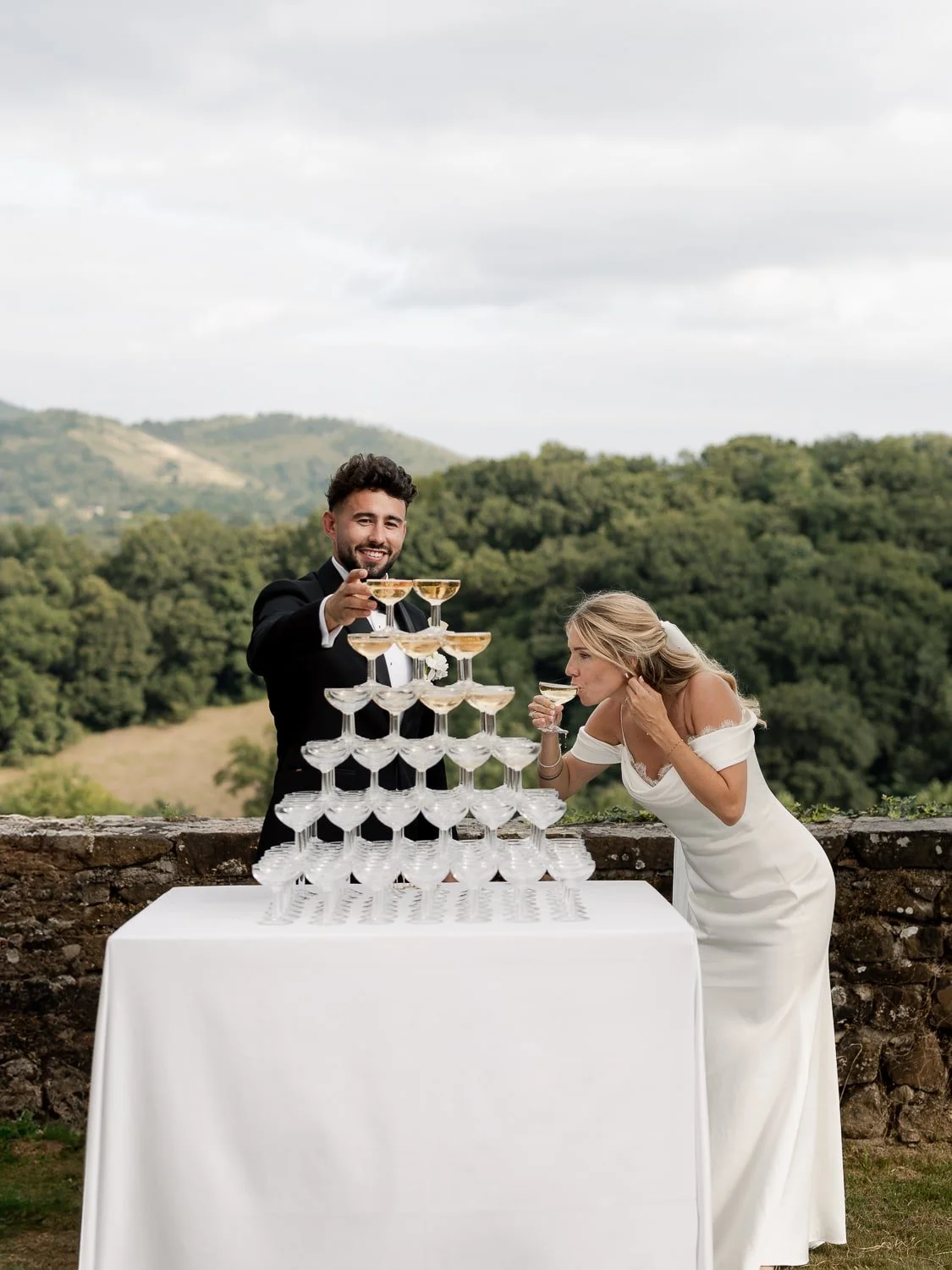 A man and woman celebrating outdoors with a champagne tower, woman in a white dress drinking from a glass.