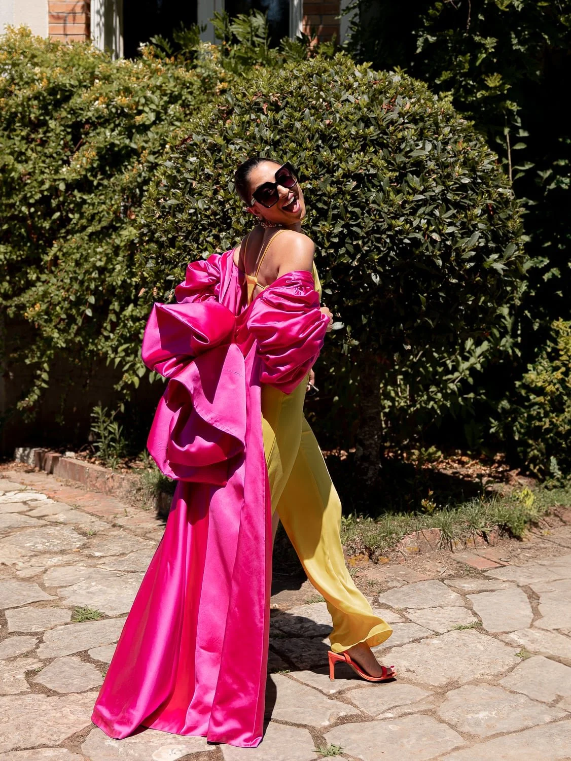 Woman wearing oversized pink coat and yellow pants with sunglasses, standing on stone pavement.
