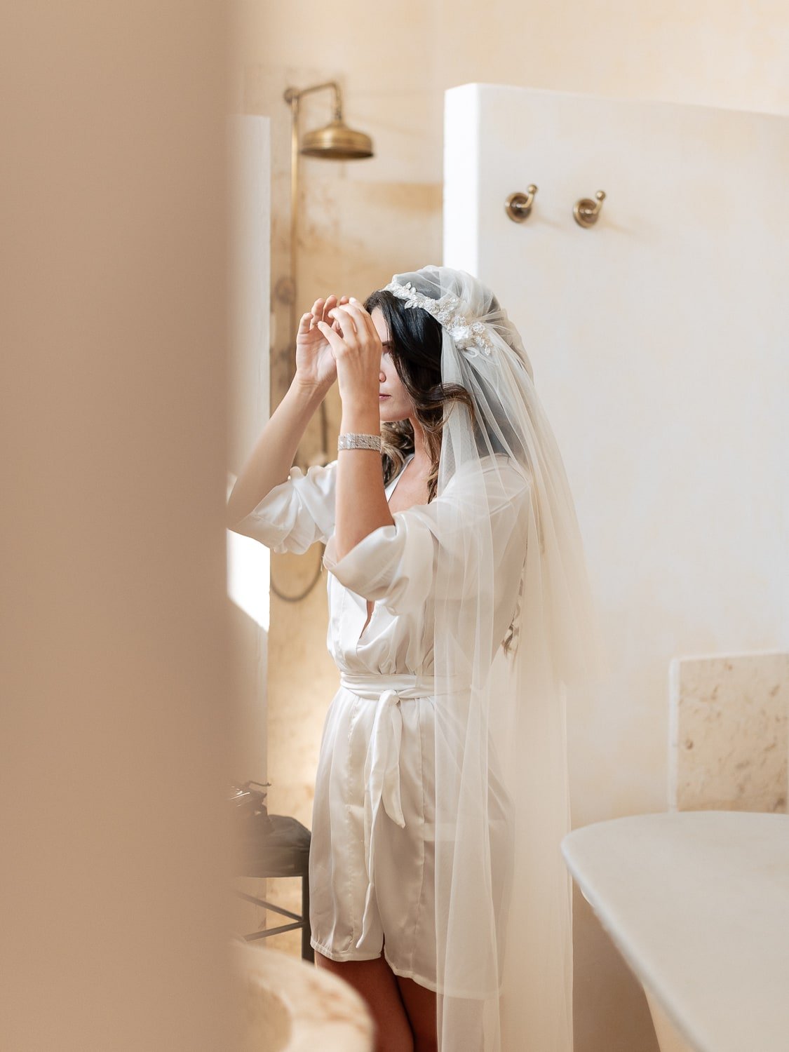 Woman in a bathroom wearing a wedding veil and robe, adjusting her hair near a mirror.