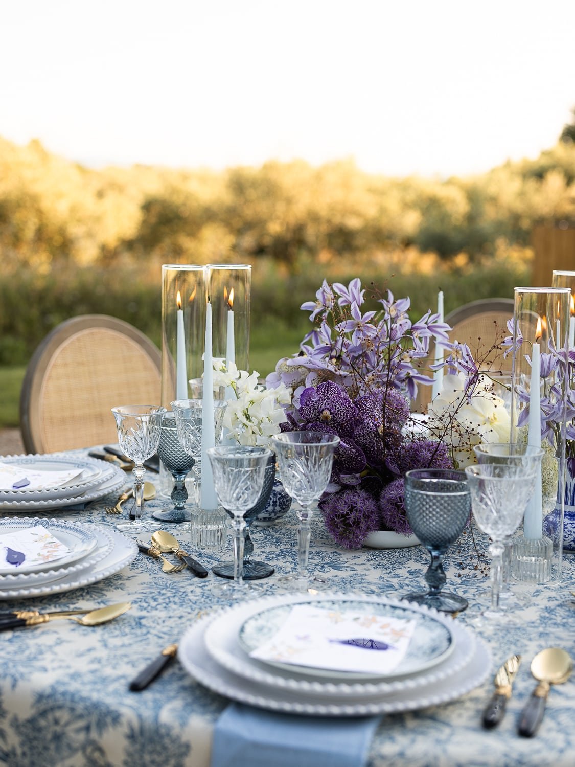 Elegant outdoor dining table with floral centerpiece, candles, glassware, and gold cutlery, set in a garden during sunset.