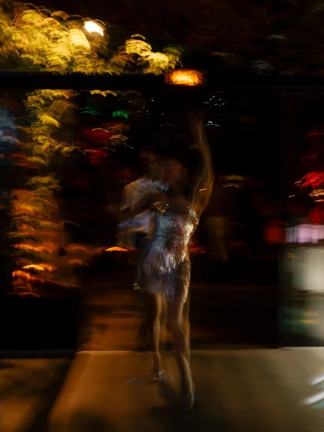 Blurred abstract image of a dancing person in a sparkling dress at night with colorful lights in the background.