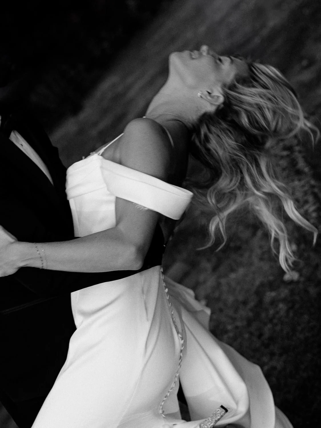 Black and white photo of a woman in a wedding dress, leaning back gracefully, with long flowing hair, held by someone in a dark suit.