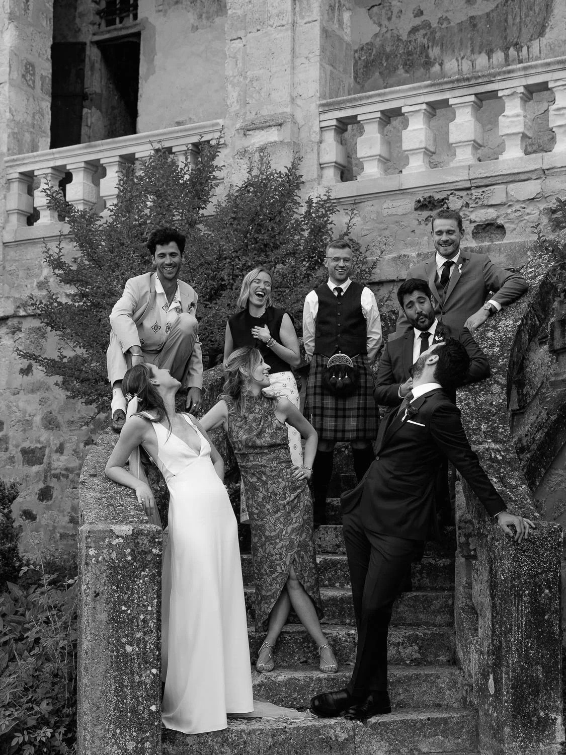 Group of people in formal attire posing playfully on stone stairs of an old building.