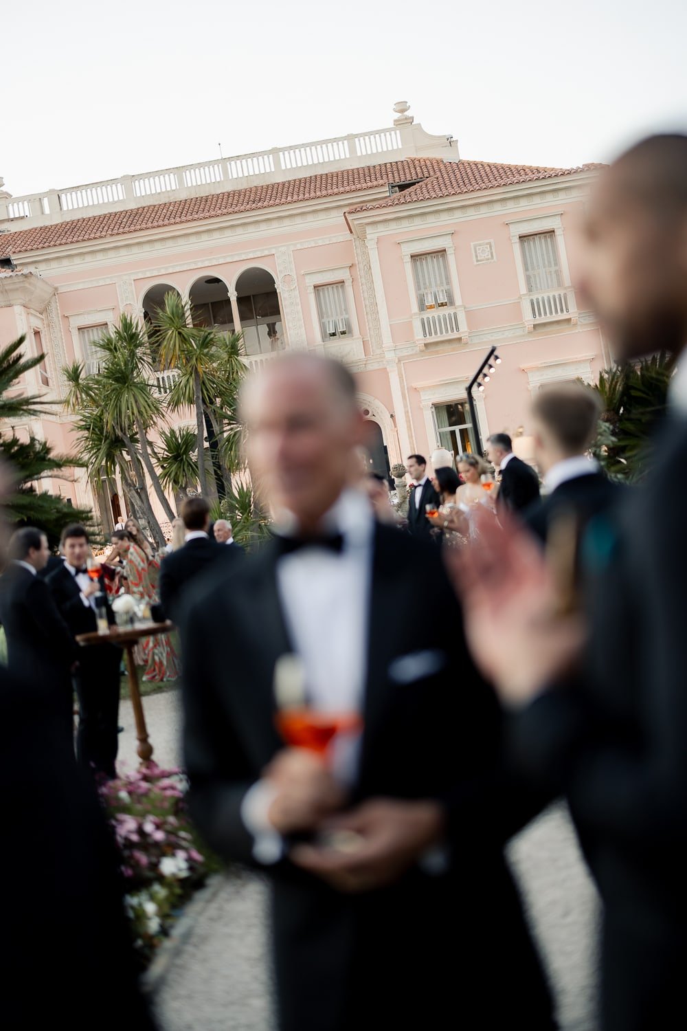 Blurry image of a formal outdoor event with several people in tuxedos, a pink mansion building with balconies, palm trees, and flowers in the background.