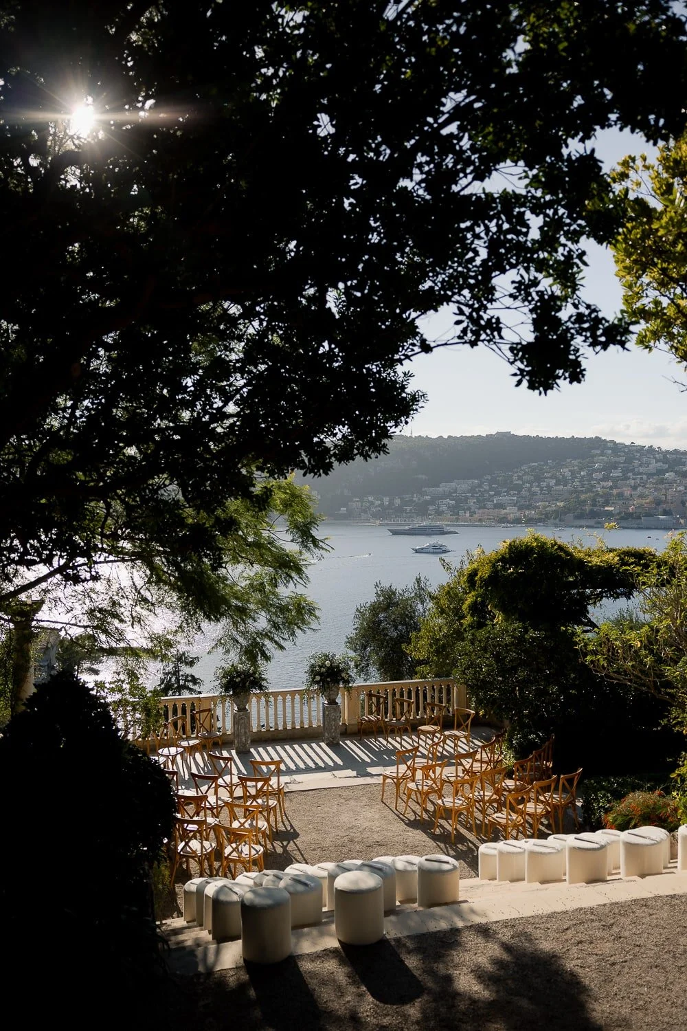 An outdoor event space with wooden chairs arranged on a gravel surface, overlooking a body of water with boats, trees, and a hillside in the background. The scene is framed by leafy branches and sunlight filtering through the trees.