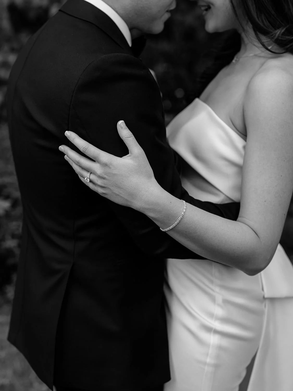 Black and white close-up of a couple embracing, wearing formal attire. The woman wears an off-shoulder dress with a bracelet and engagement ring.