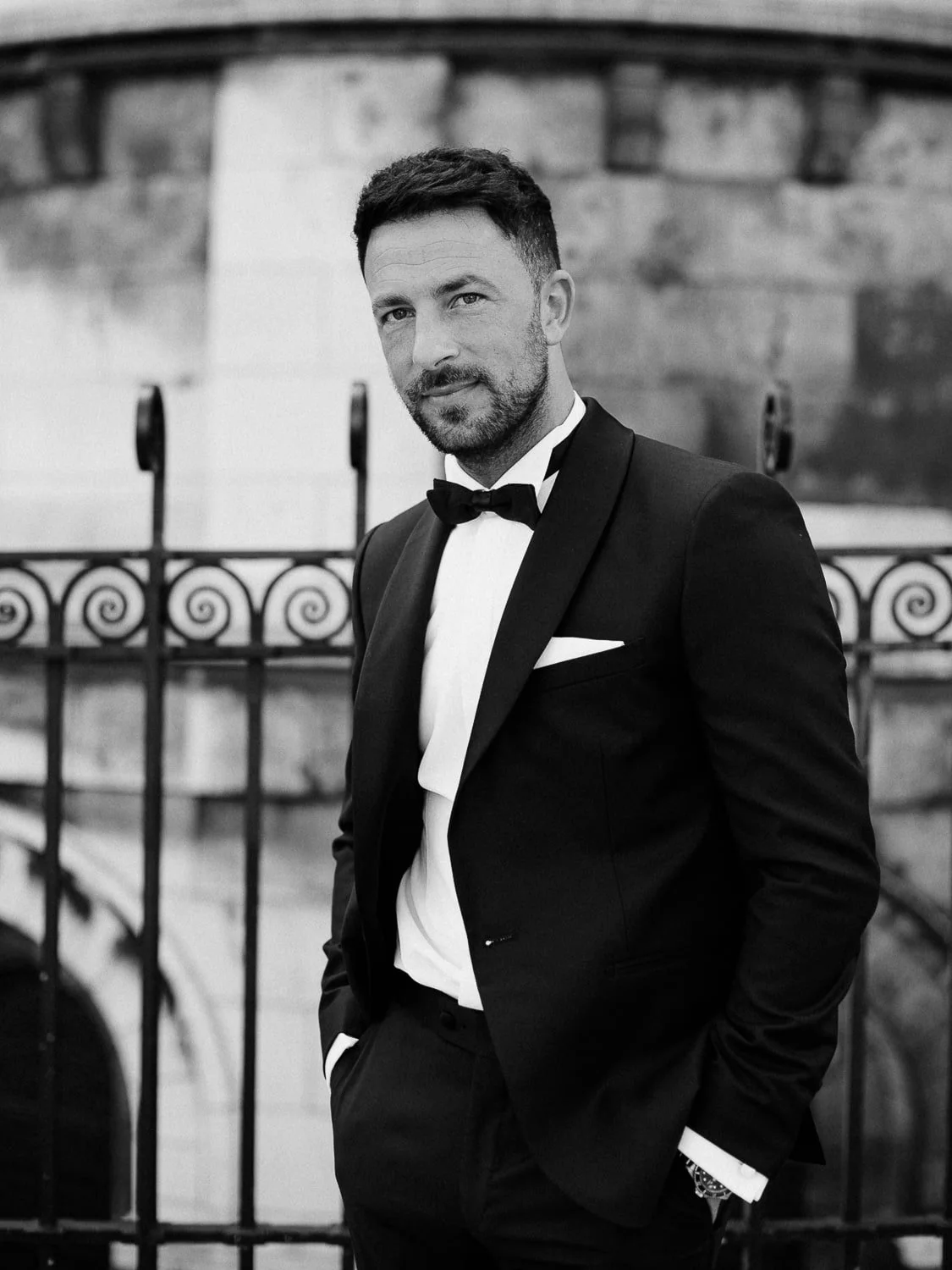Man in a suit with a bow tie standing outdoors in front of a decorative metal fence, black and white image.