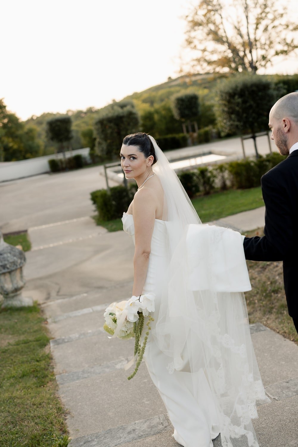 Bride in white wedding dress holding a bouquet, with a veil, walking outdoors with man in black suit, trees and mountains in the background at sunset.