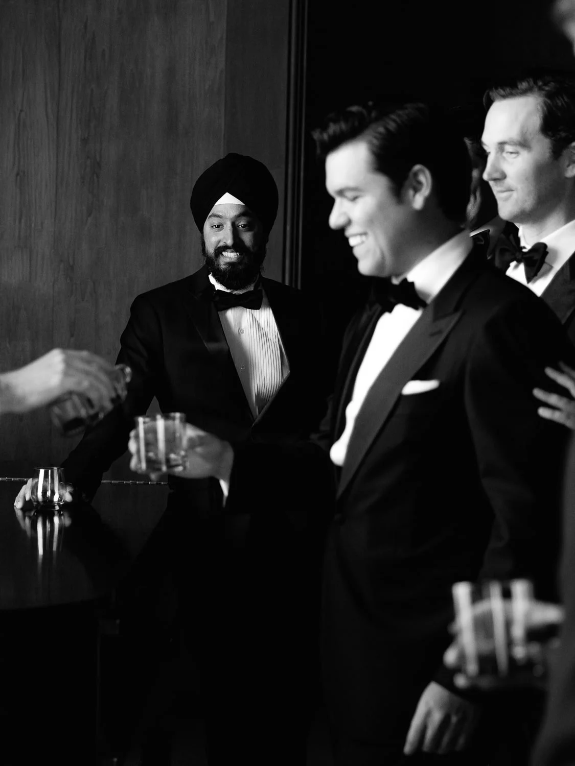 Three men in tuxedos at a formal event, smiling, with one man wearing a turban and drinking a glass of liquor, in a black and white photograph.