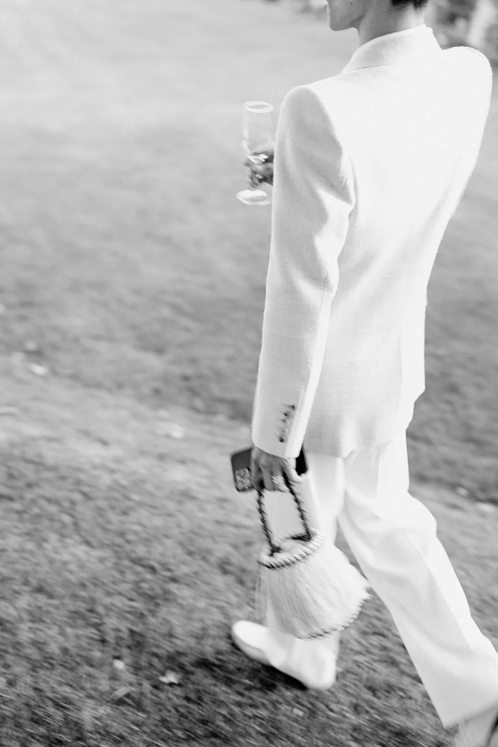 Person in white suit holding a wine glass and a handbag, walking on grass.