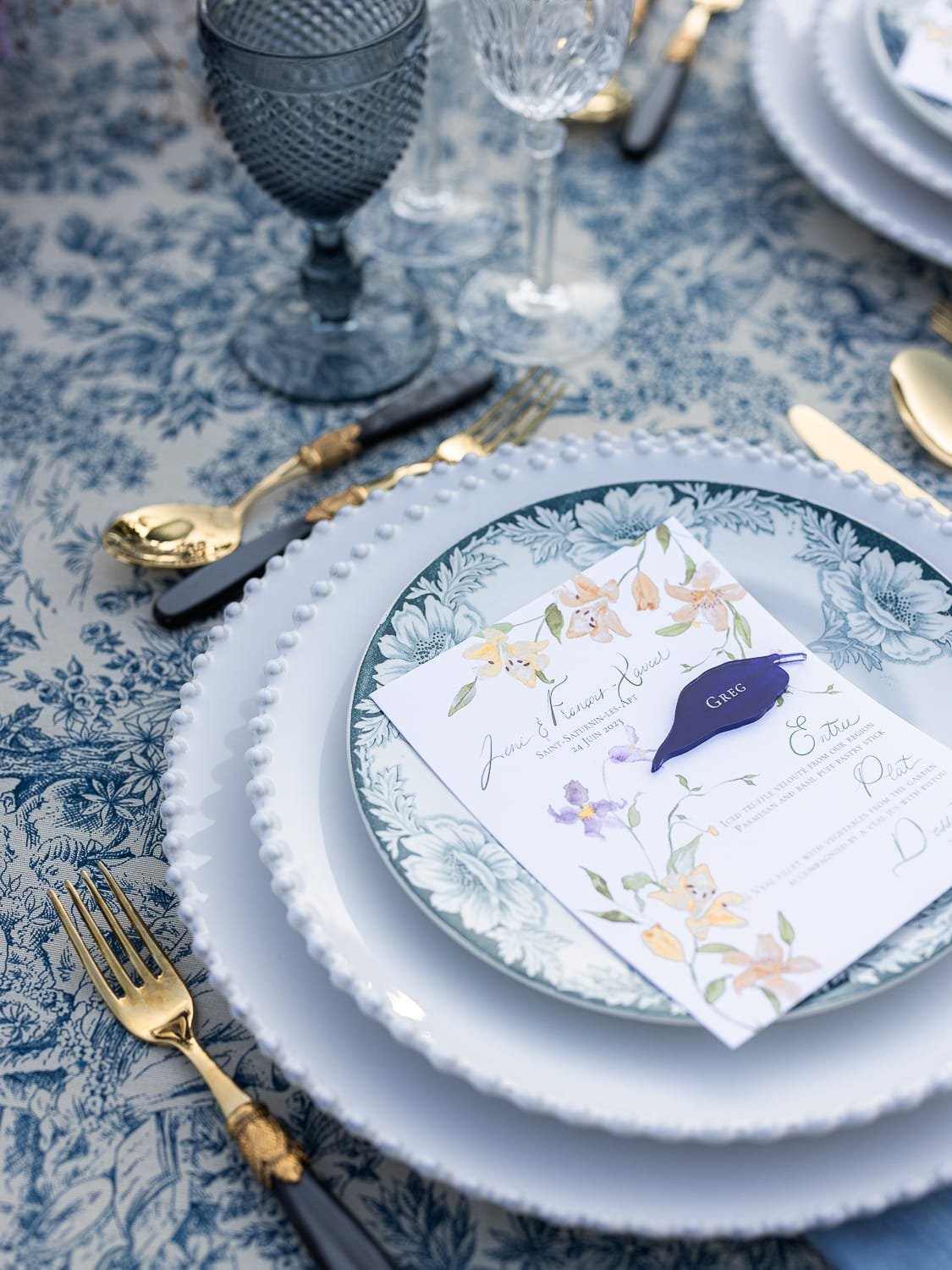Elegant table setting with blue floral tablecloth, layered decorative plates, and gold cutlery. Glasses and a floral-printed menu card with a name tag are placed on the plates.