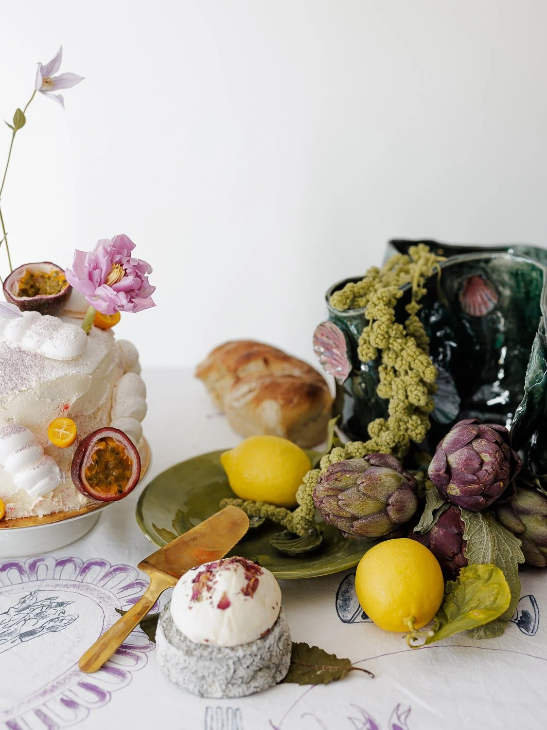 Artistic food display with a decorated cake featuring flowers and passion fruit, a loaf of bread, a green plate with artichokes, lemons, and green flowers, and a dessert with rose petals on a granite-like base, set on a table with a patterned cloth.