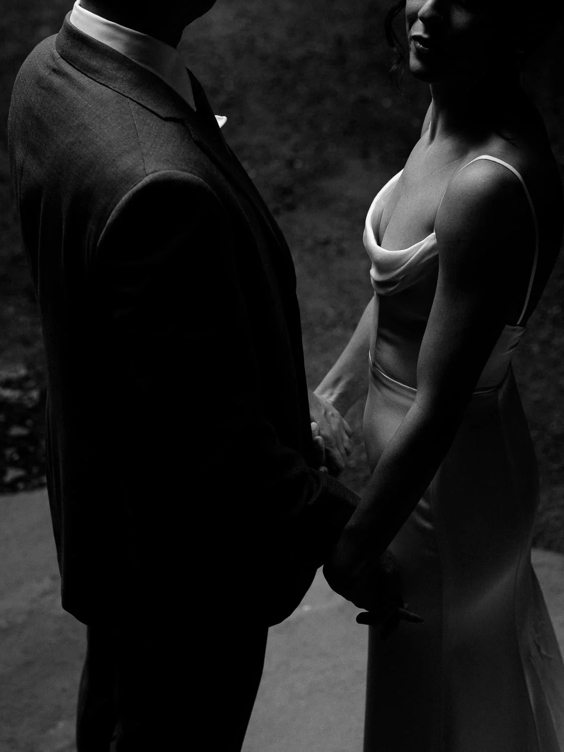 Black and white photo of a couple holding hands, featuring a man in a suit and a woman in an elegant satin dress, captured from the waist up.