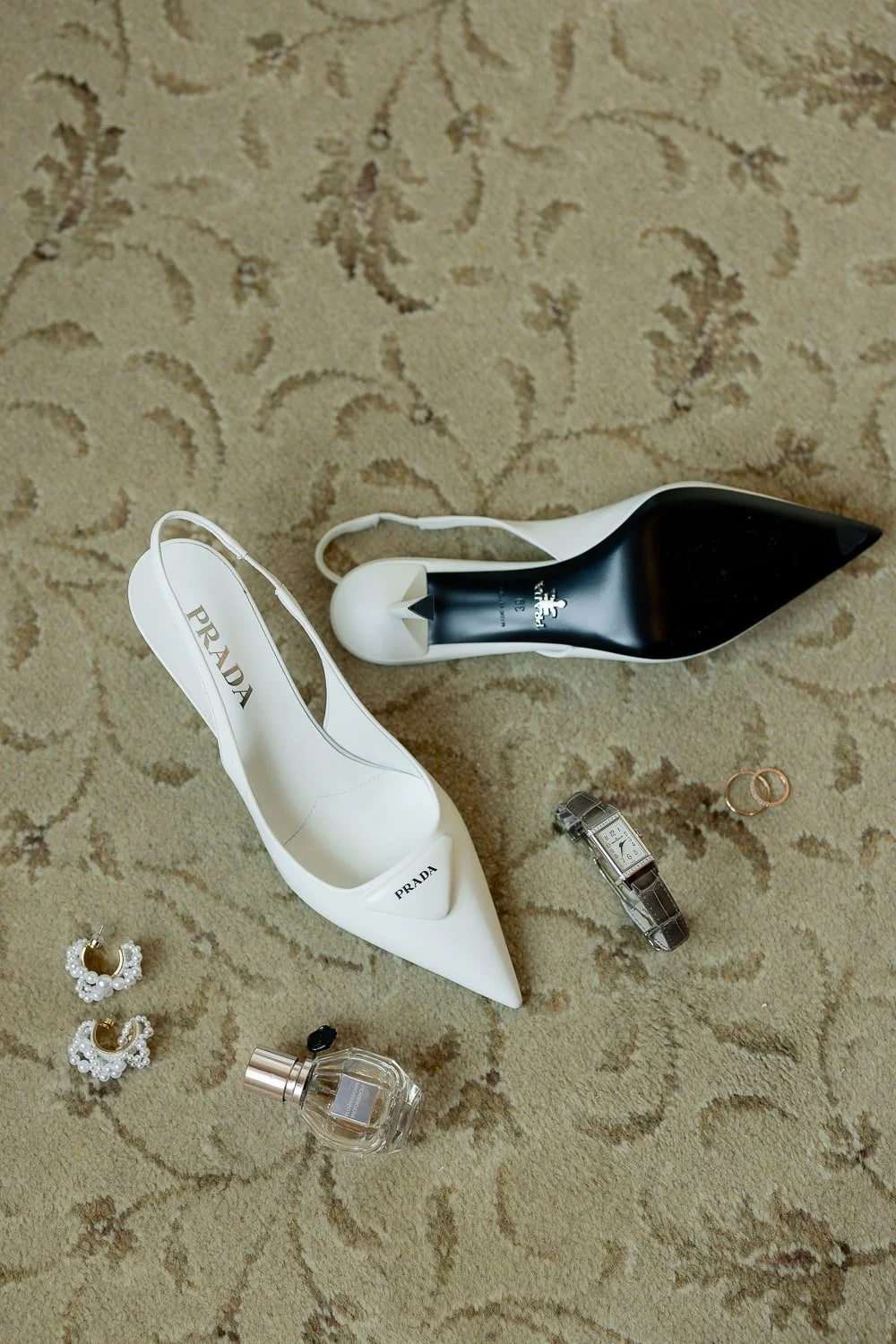 White Prada slingback heels, silver watch, two rings, pearl earrings, and perfume bottle on a patterned carpet.