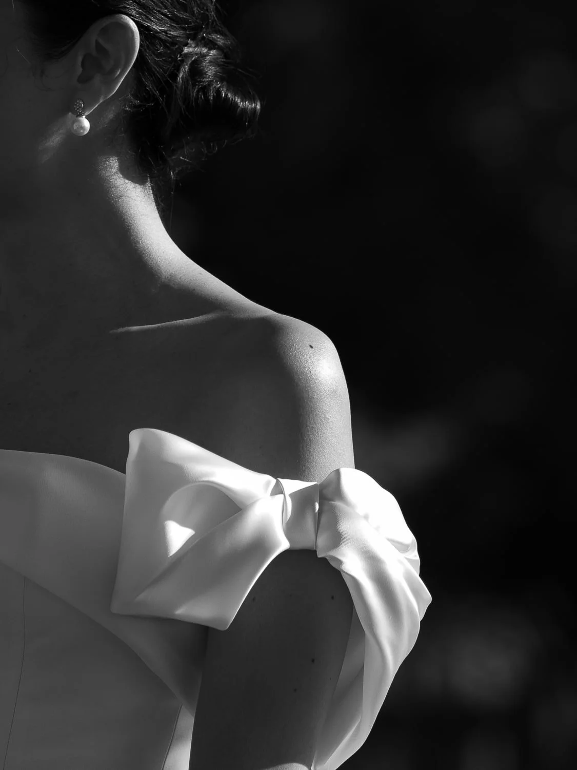 Black and white photo of a woman wearing an off-the-shoulder dress with a large bow on the sleeve, pearl earring visible, hair is styled up.