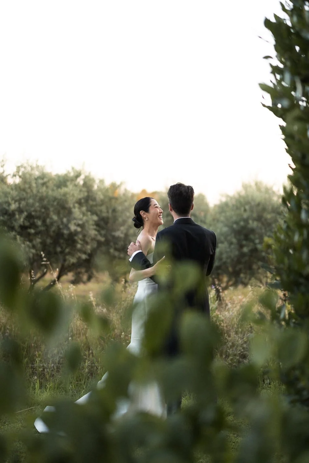 Bride and groom embracing outdoors, surrounded by trees and greenery.