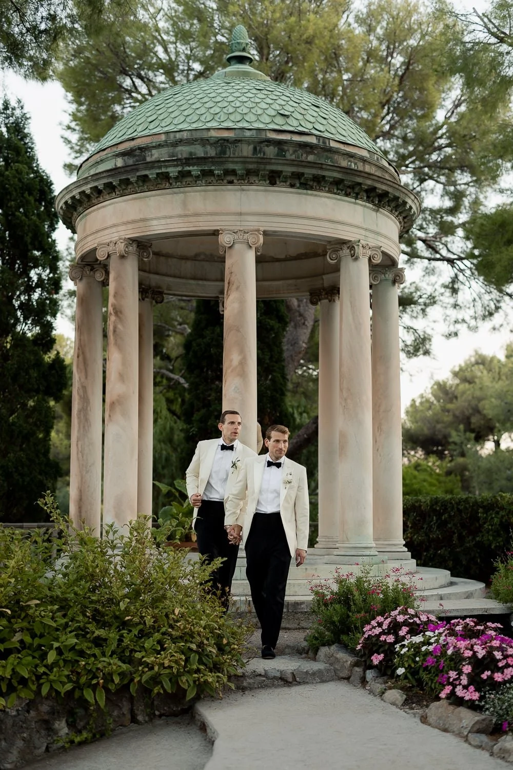 Two men dressed in tuxedos, holding hands, walking in a garden with a classical pavilion in the background.