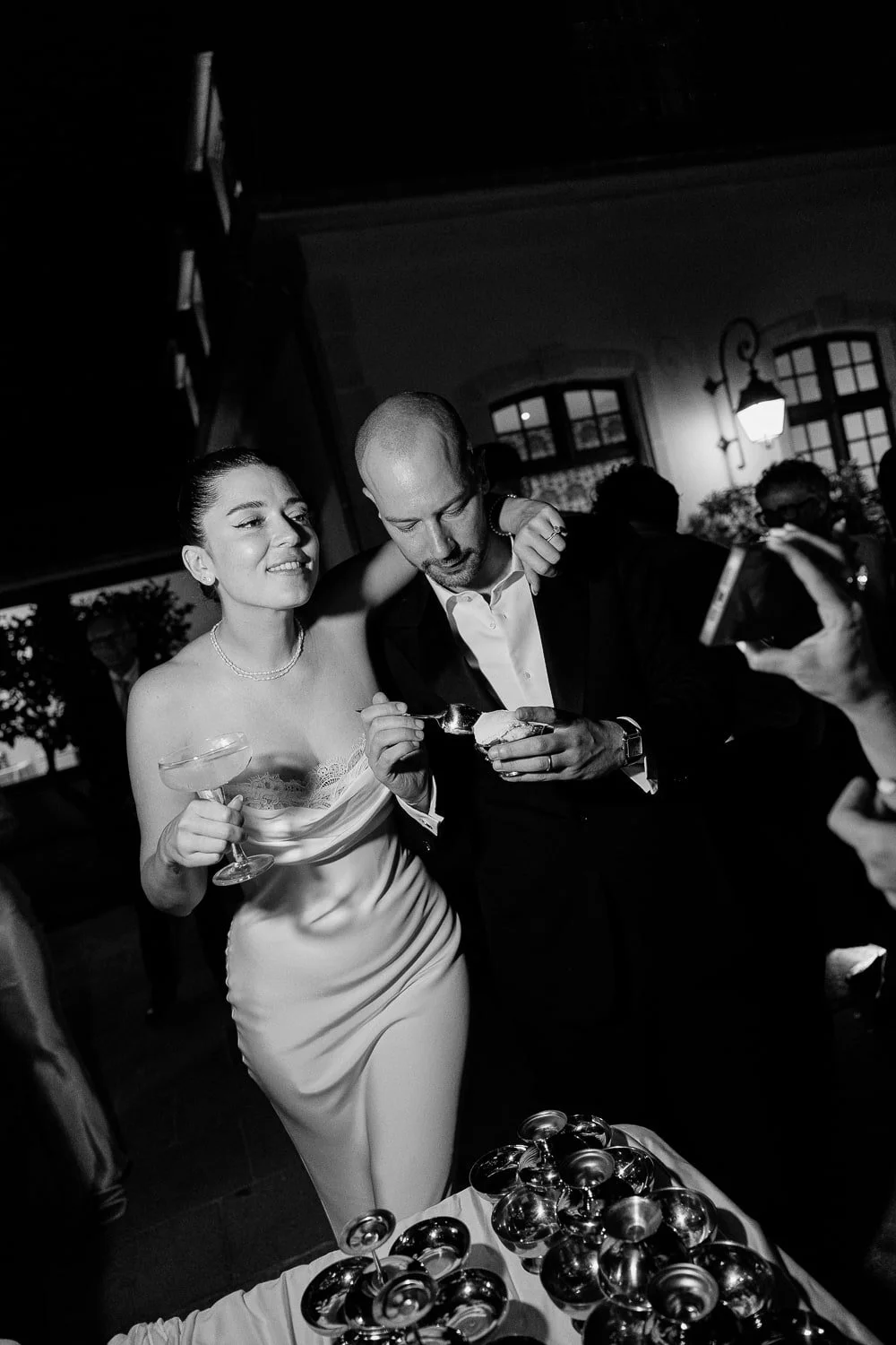 A woman in a white dress and a man in a suit sharing a dessert at a celebration, surrounded by people and a table with multiple bowls.