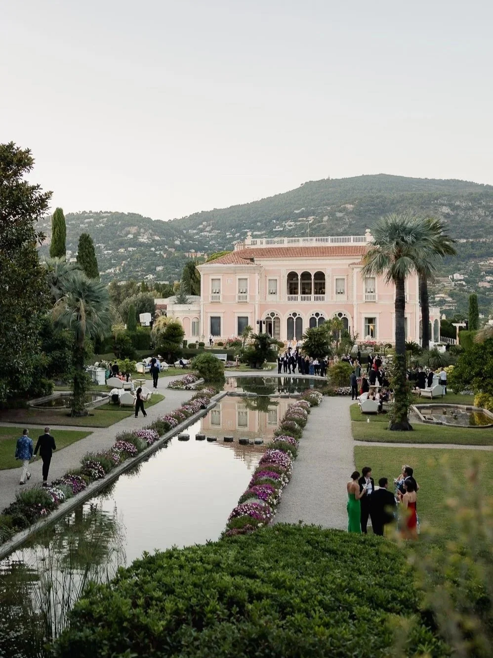 A large pink mansion surrounded by well-maintained gardens, with a pathway, palm trees, and people gathered at an outdoor event.
