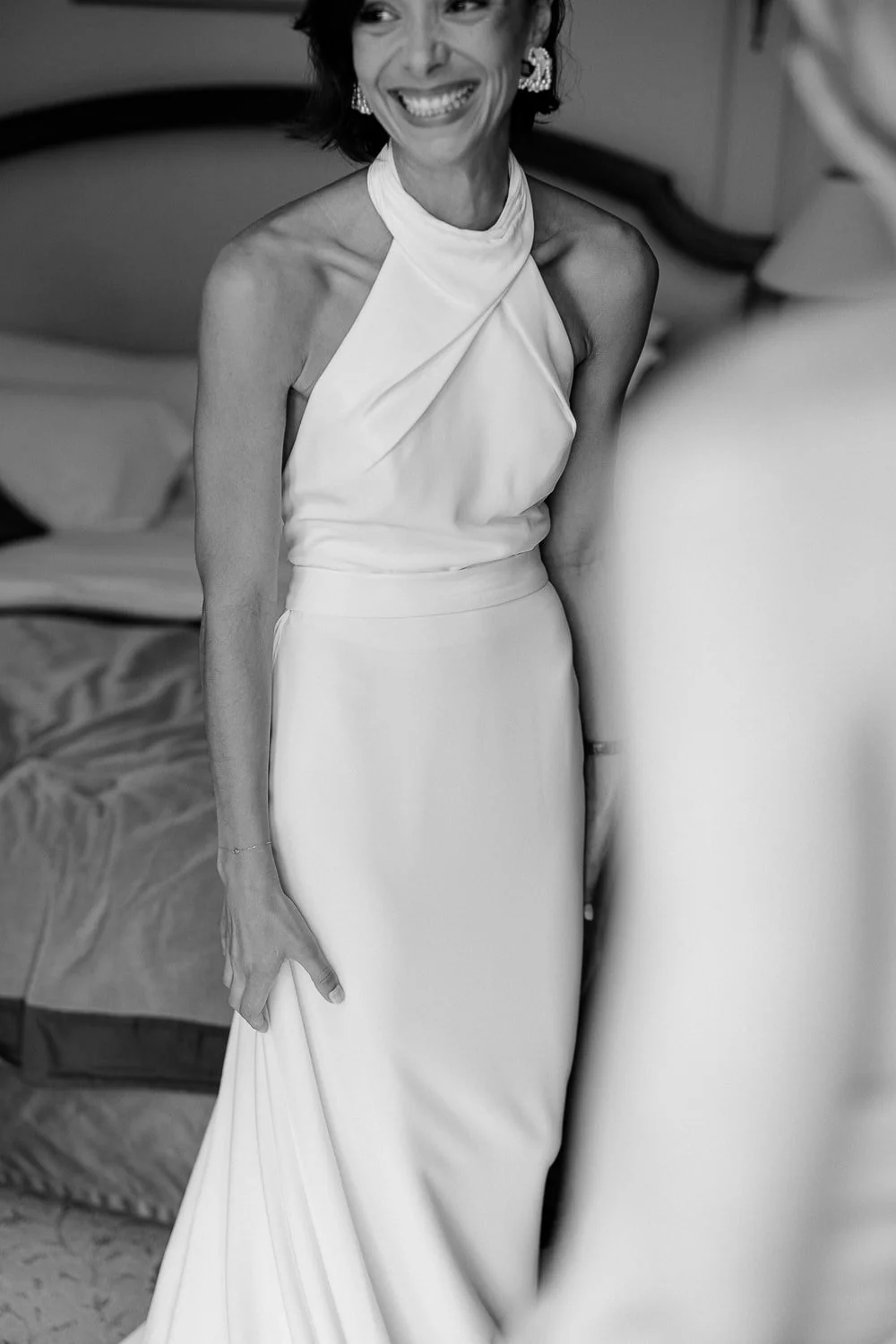 Black and white photo of a smiling woman in an elegant halter neck dress, standing indoors.