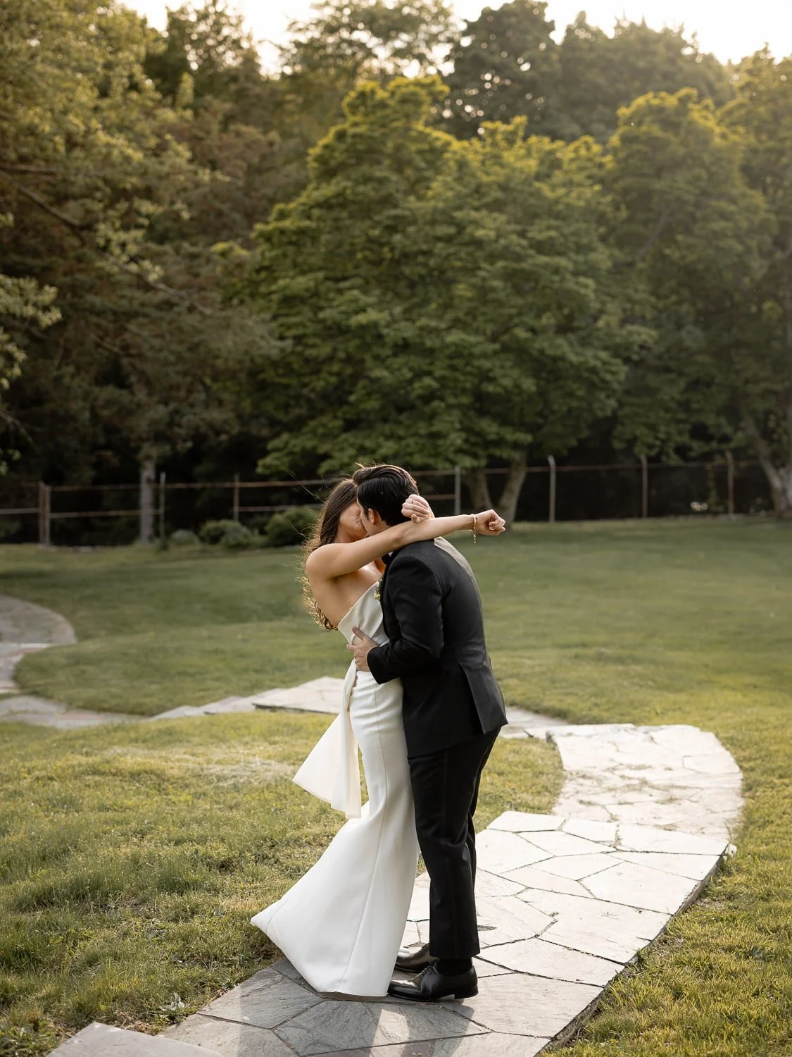 A couple hugging on a stone path in a garden, with the woman wearing a white dress and the man in a black suit, surrounded by trees.
