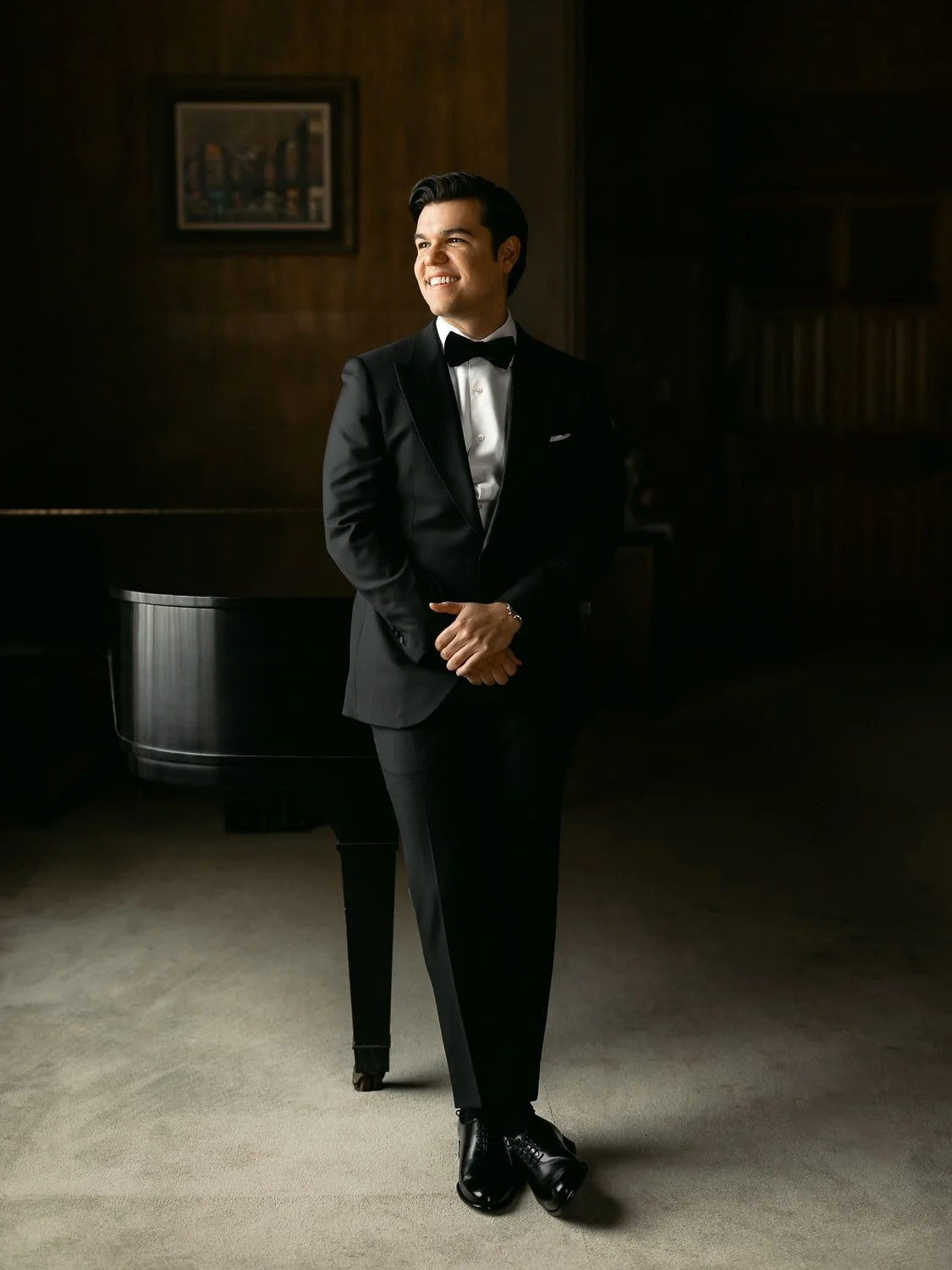 Man in formalwear standing by a grand piano in a dimly lit room.