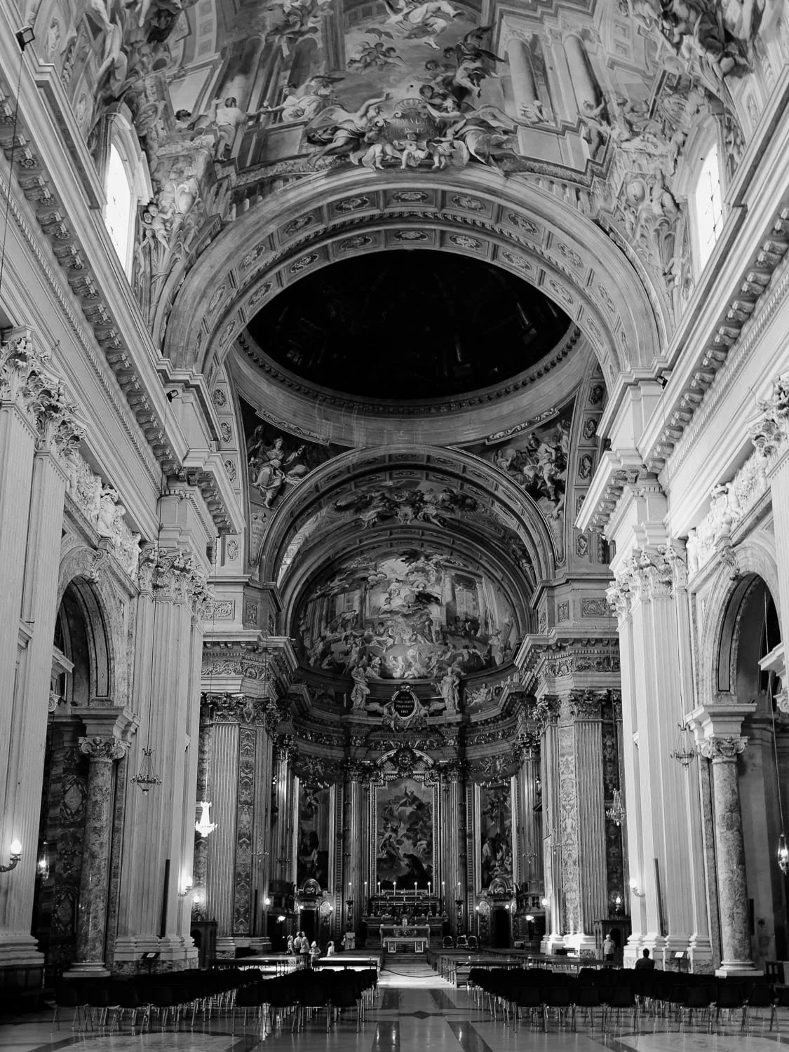 Interior of a grand cathedral with intricate Baroque architecture, high ceilings, and detailed frescoes. The image is in black and white, showcasing tall columns, ornate arches, and an altar at the center.
