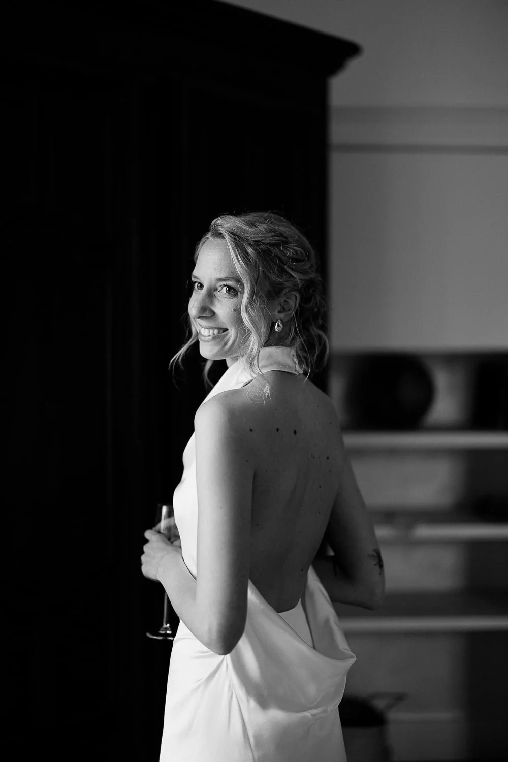 Smiling woman in a white backless dress holding a champagne glass, black and white photo.