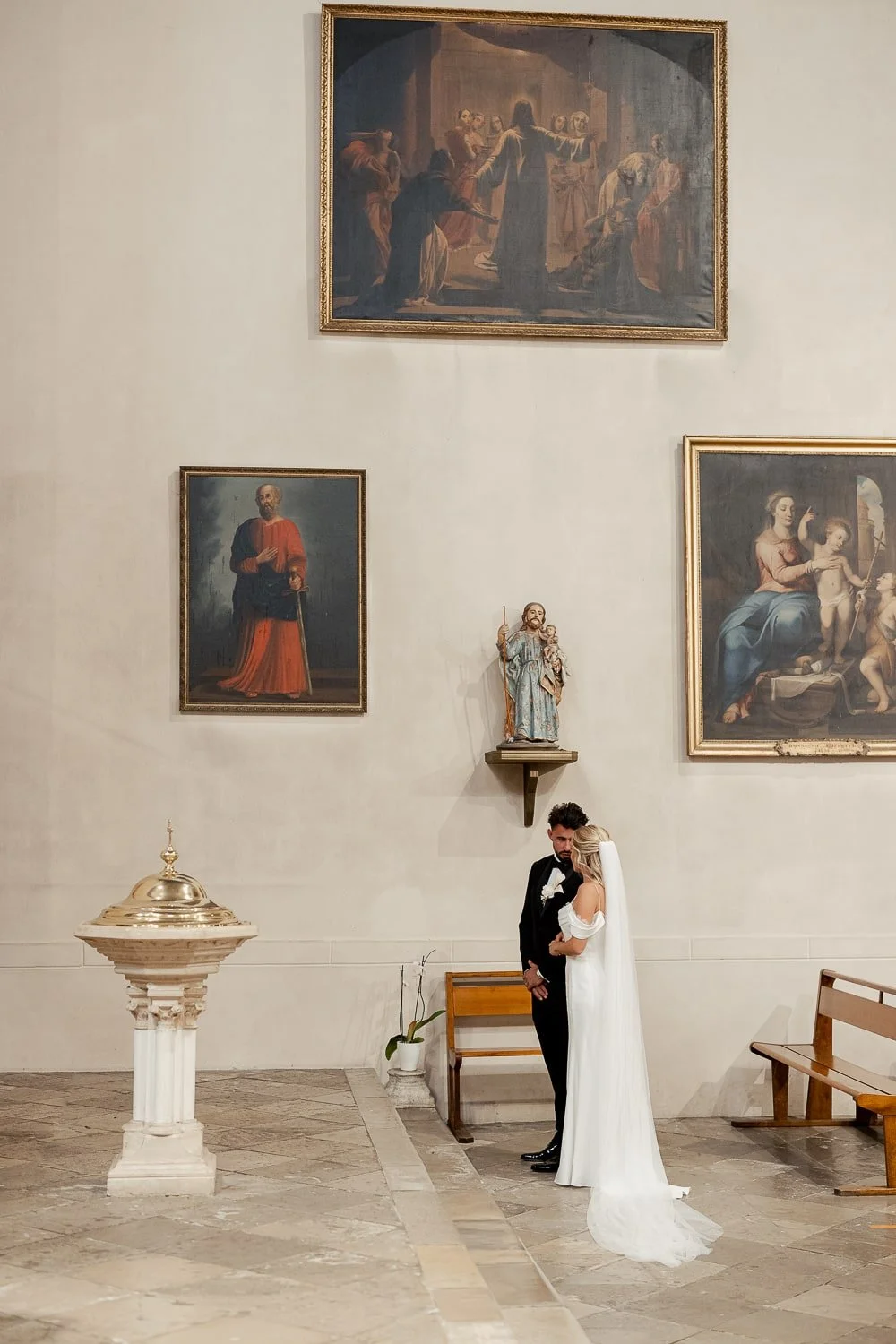 Bride and groom standing in a church with religious paintings and statue.