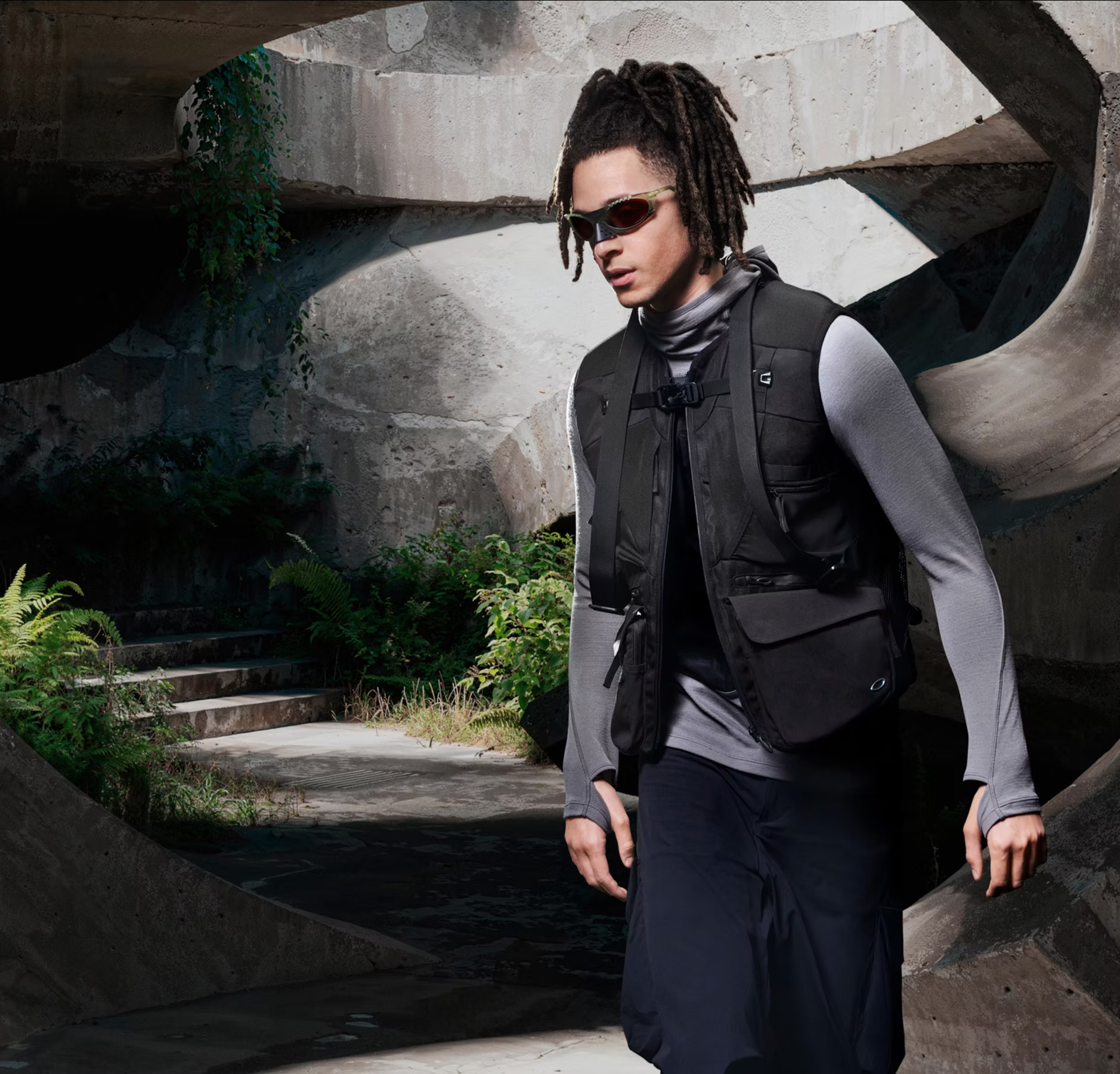 A person with dreadlocks wearing sunglasses, a gray long-sleeve shirt, and a black vest, carrying a backpack, standing outdoors near concrete and greenery.