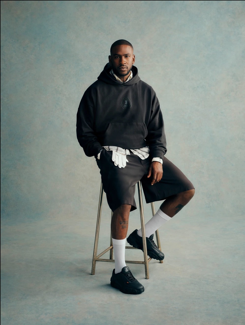 A man wearing a black hoodie, shorts, white socks, black sneakers, and gloves, sitting on a stool against a plain background.
