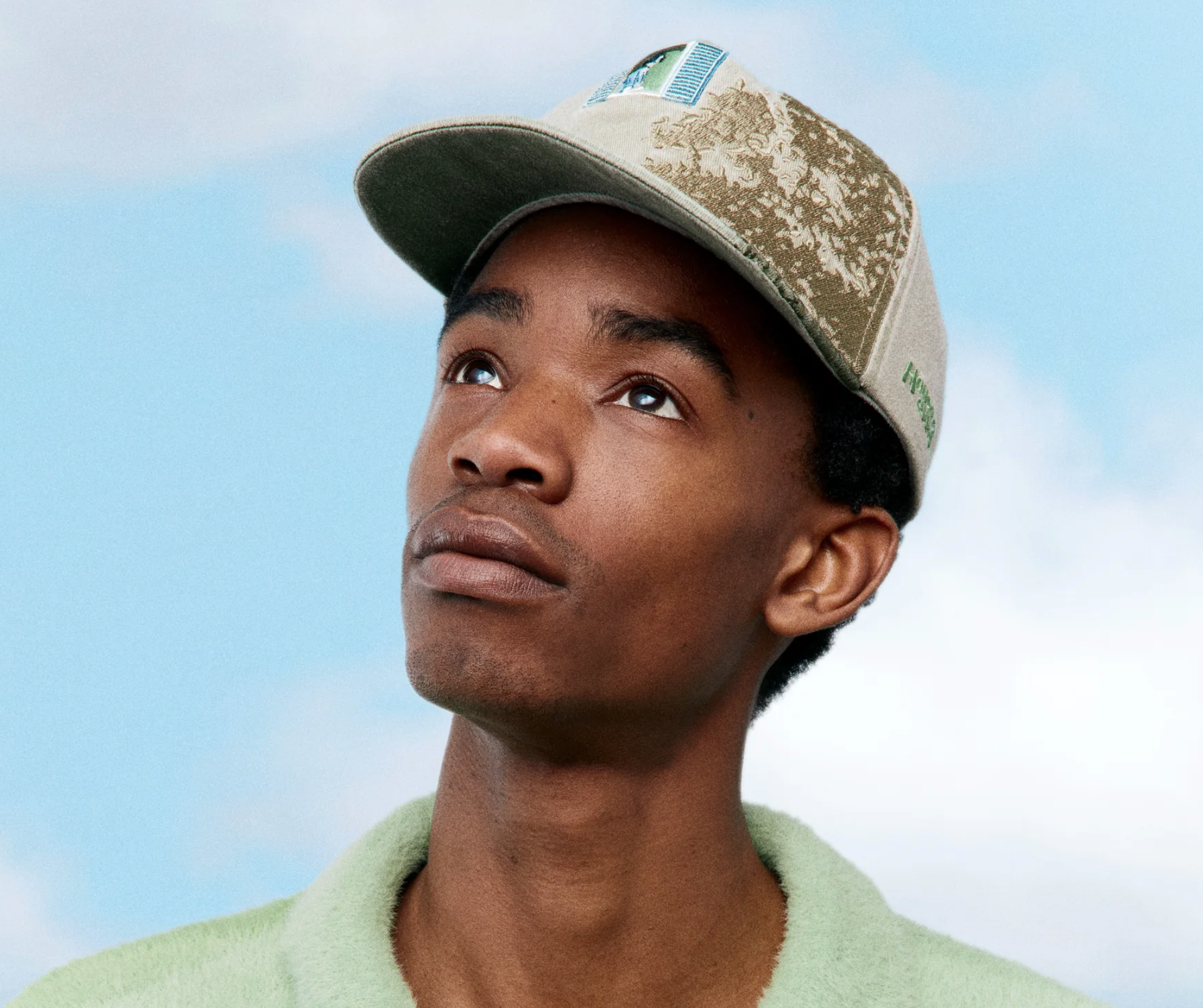 A young man with dark skin looking up while wearing a beige cap with a landscape pattern and a green sweater against a cloudy sky.