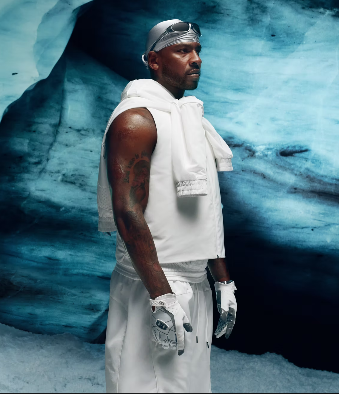 A man dressed in white athletic gear, including gloves, with sunglasses on his head, standing in front of an icy background, possibly at a cold environment or ice rink.