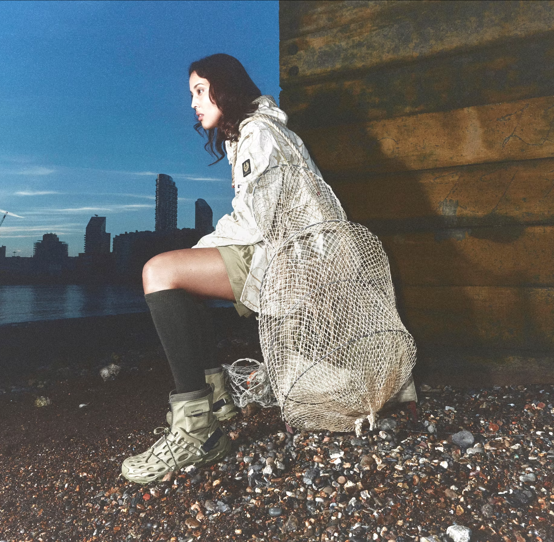 A woman sitting on a rocky shoreline at dusk, holding a fishing net, with a city skyline and river in the background, and a wooden wall on her right.