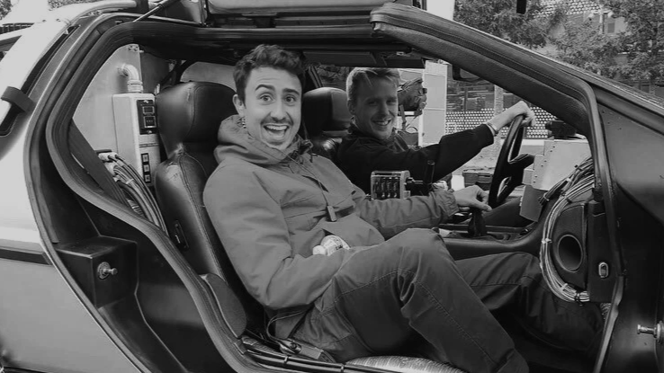 Two smiling men sitting inside a futuristic car with a partially open door, with one man in the driver's seat and the other in the passenger seat, both looking at the camera.