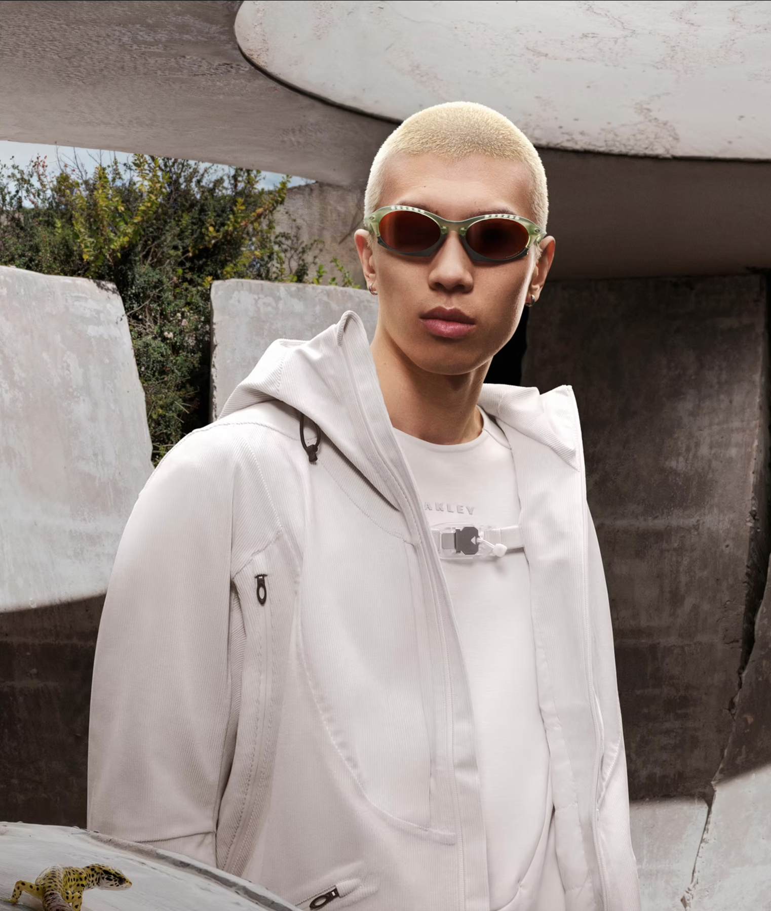 Person with a buzz cut hairstyle wearing sunglasses, a white jacket, and a white shirt standing outdoors near concrete structures.