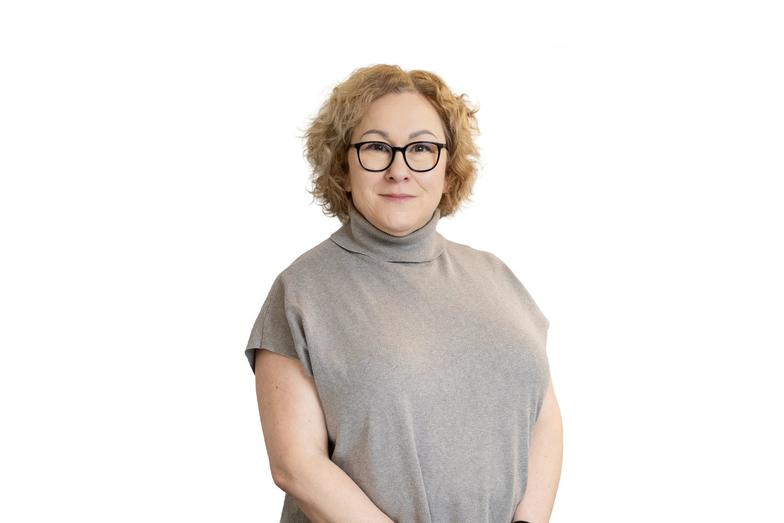 A middle-aged woman with curly blonde hair, wearing glasses and a beige turtleneck shirt, standing against a white background.