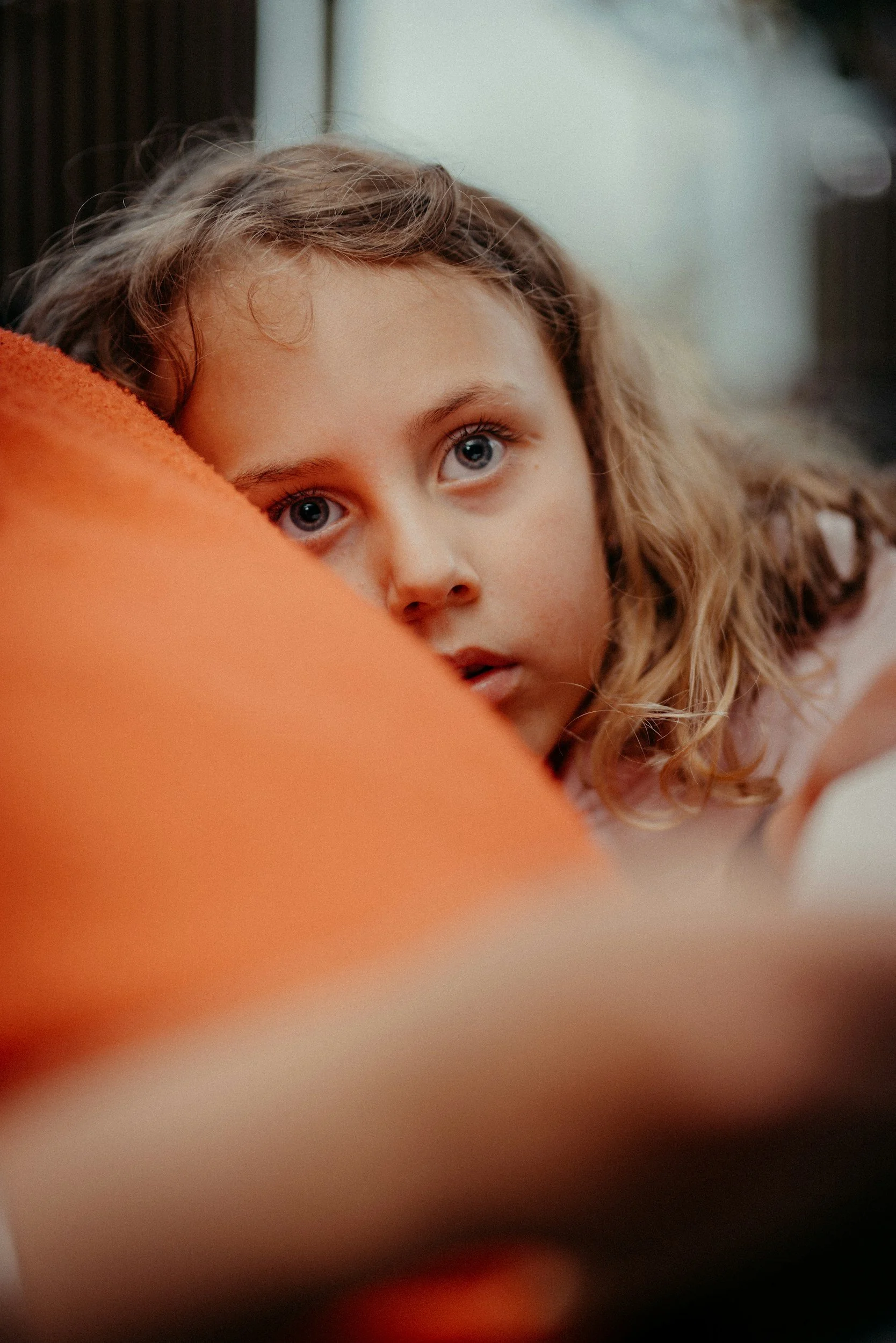 Una niña con cabello rubio y ojos azules, descansando con la cabeza apoyada en un cojín naranja, en un ambiente interior con luz natural y fondo difuso.